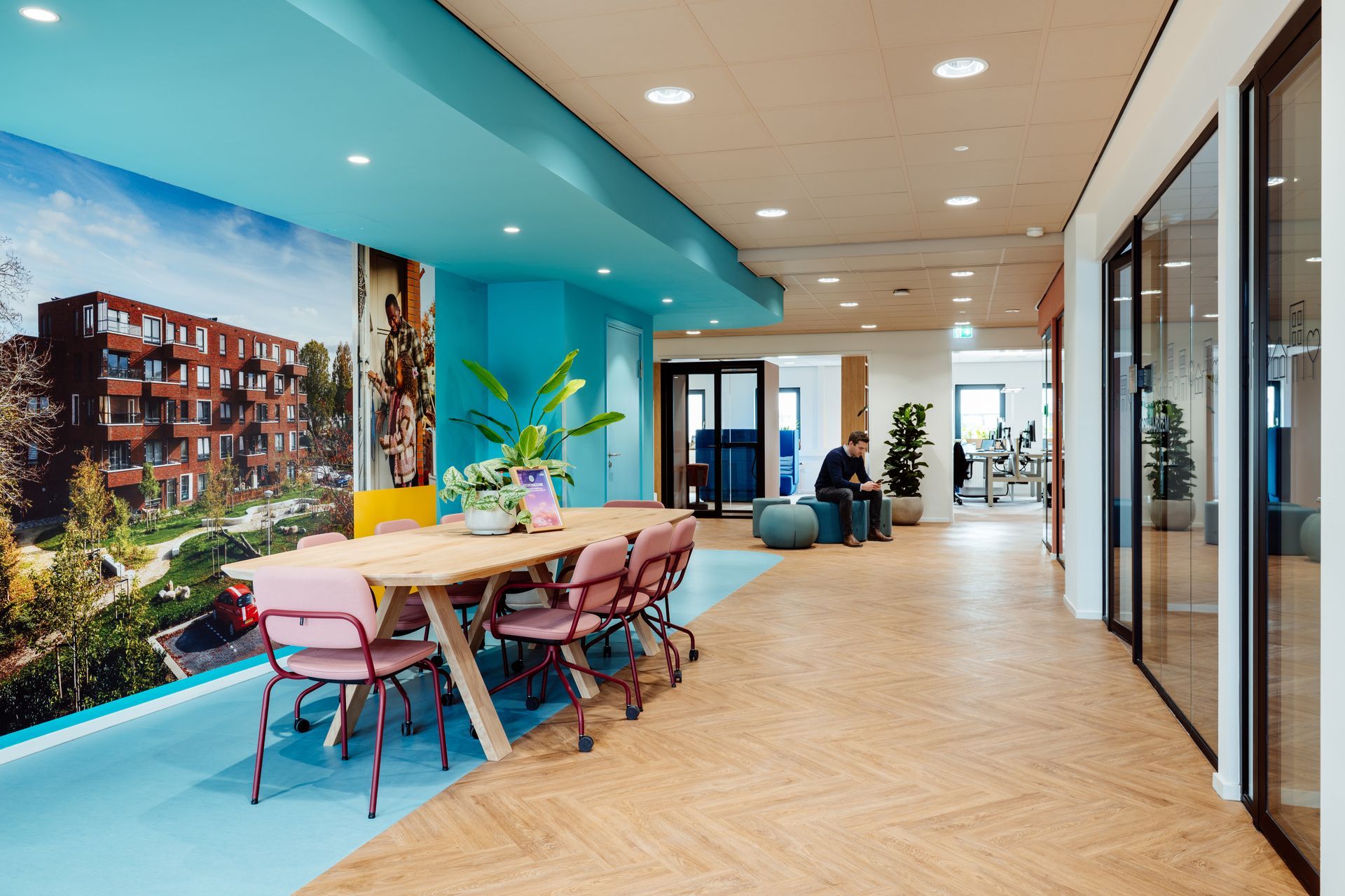 Moderne kantoorlounge met roze stoelen, houten tafels, een turquoise muur en een vergaderruimte met glazen wanden. interieurontwerp door KuóStudio