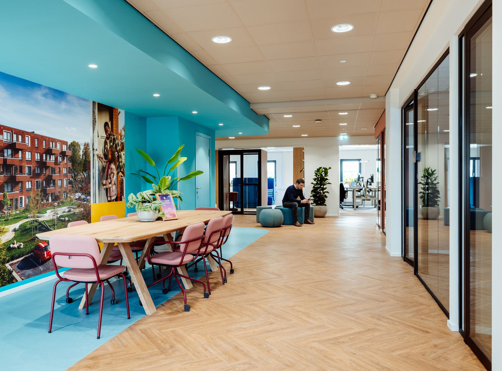 Modern office lounge with pink chairs, long table, blue ceiling, and glass-walled hallway