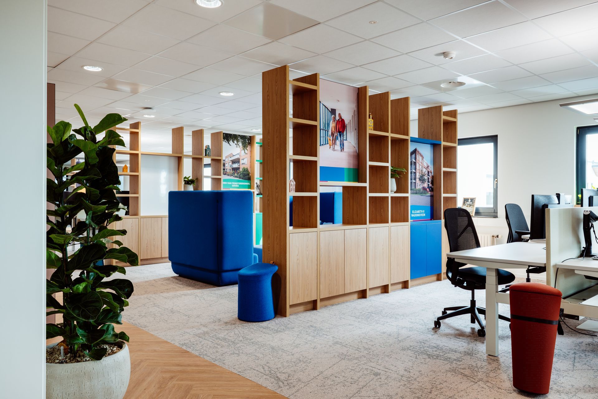 Open kantoorruimte met houten schappen, bureaus, stoelen en blauwe loungebanken onder heldere plafondverlichting.