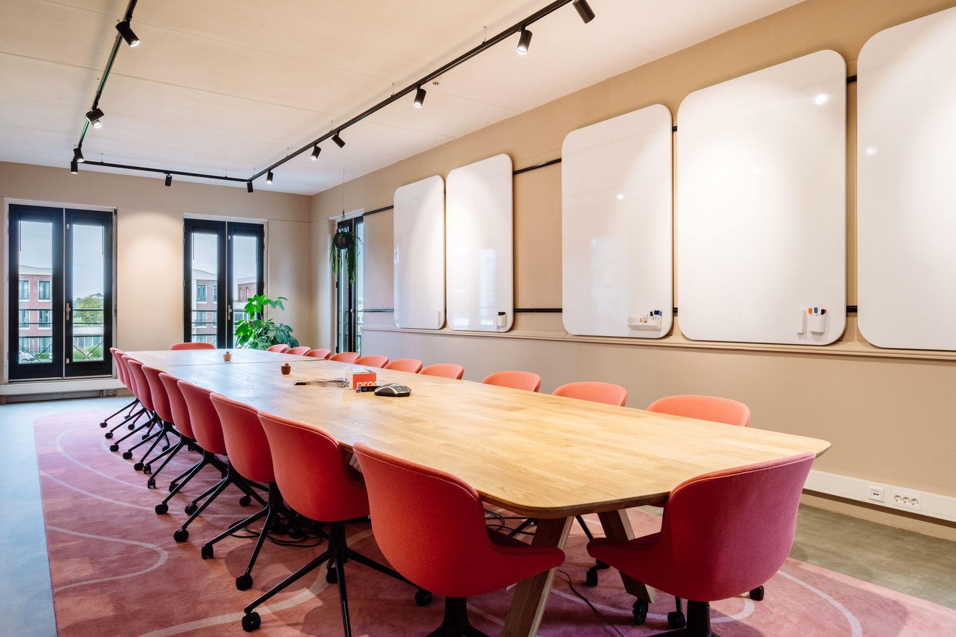 Een vergaderruimte met een lange houten tafel, rijen zalmkleurige stoelen, een roze tapijt met patroon en whiteboards aan de muur in het nieuwe kantoor van The Future Group Interieurontwerp KuóStudio