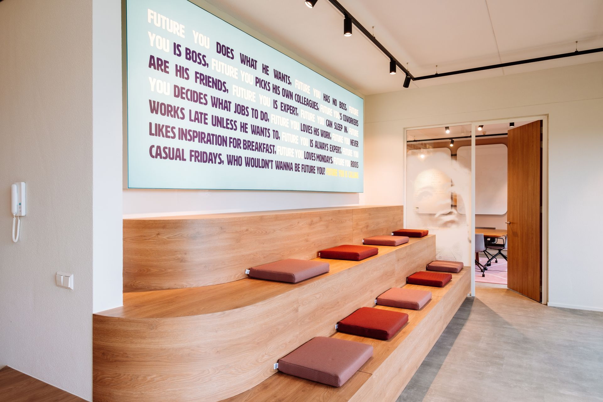 Houten tribune met kussens, geplaatst tegen een muur met een groot tekstbord in een lichte, moderne ruimte in het nieuwe kantoor van The Future Group Interieurontwerp KuóStudio