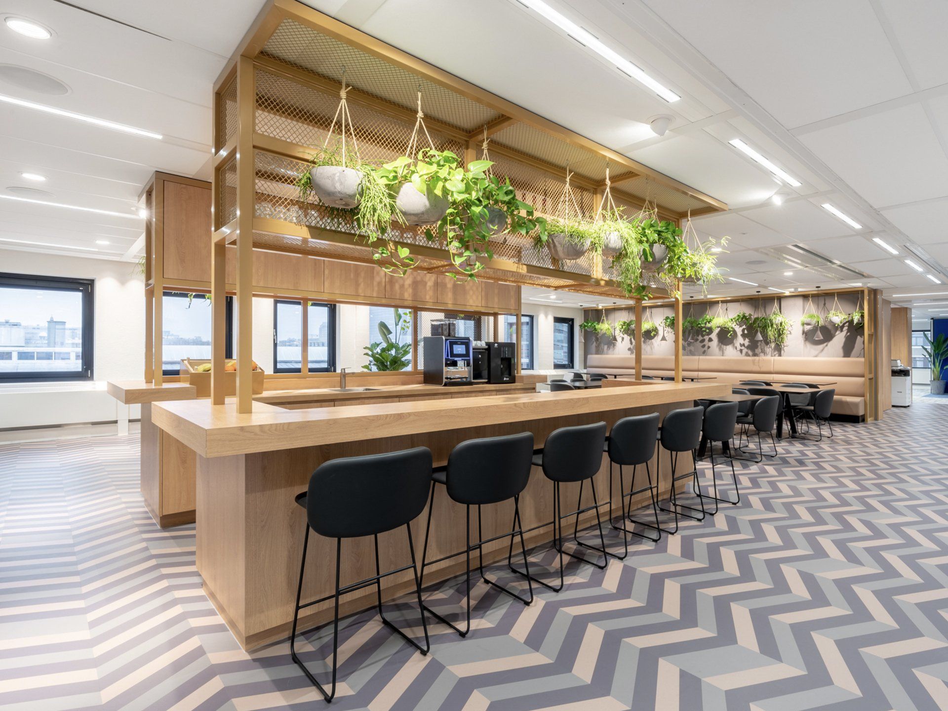 Moderne kantoorlounge met houten bar, zwarte barkrukken, hangplanten en een tapijt met zigzagpatroon.