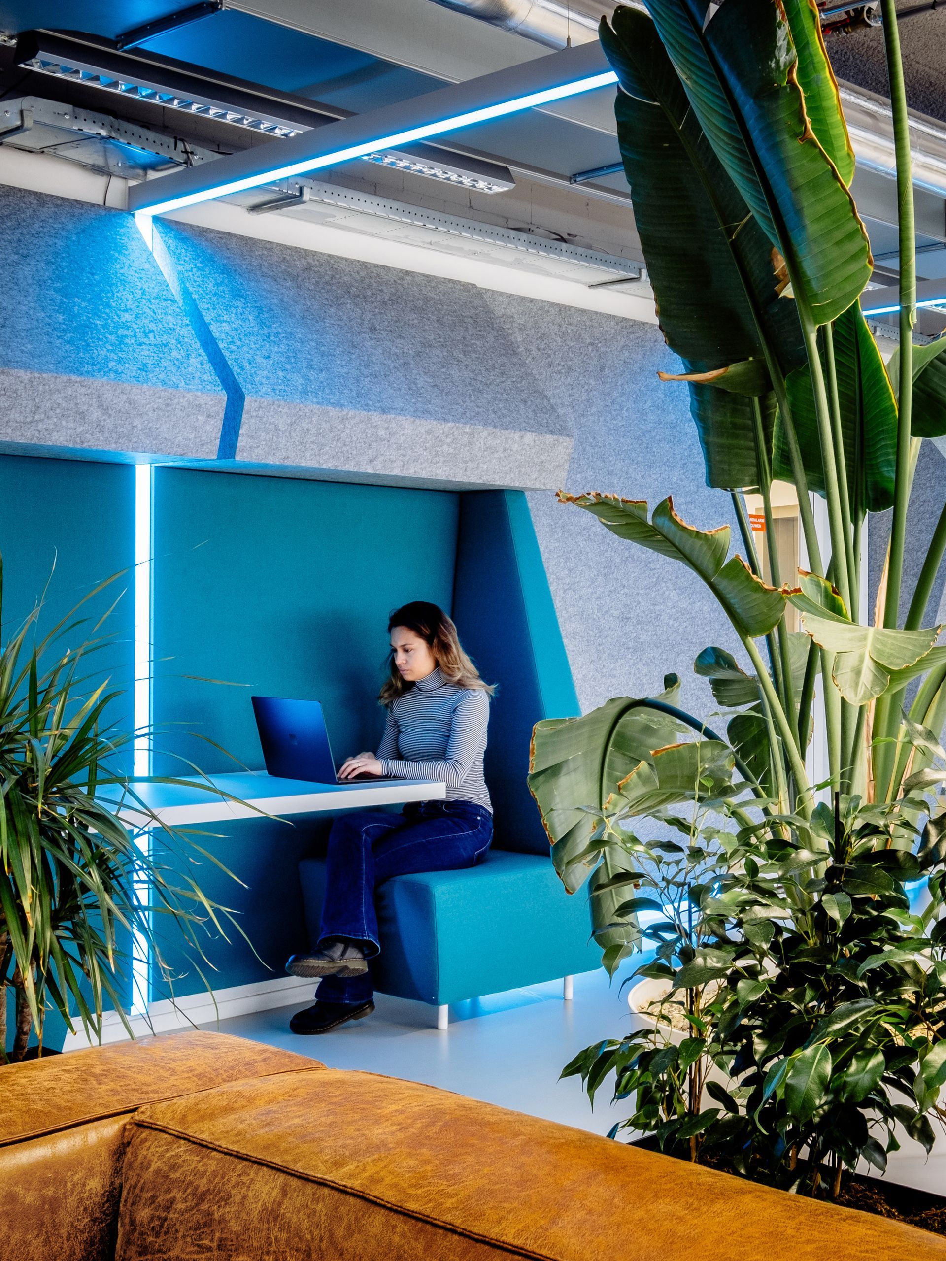 Een persoon werkt in een blauwe cabine in een moderne binnenlounge met planten en geometrische verlichting.