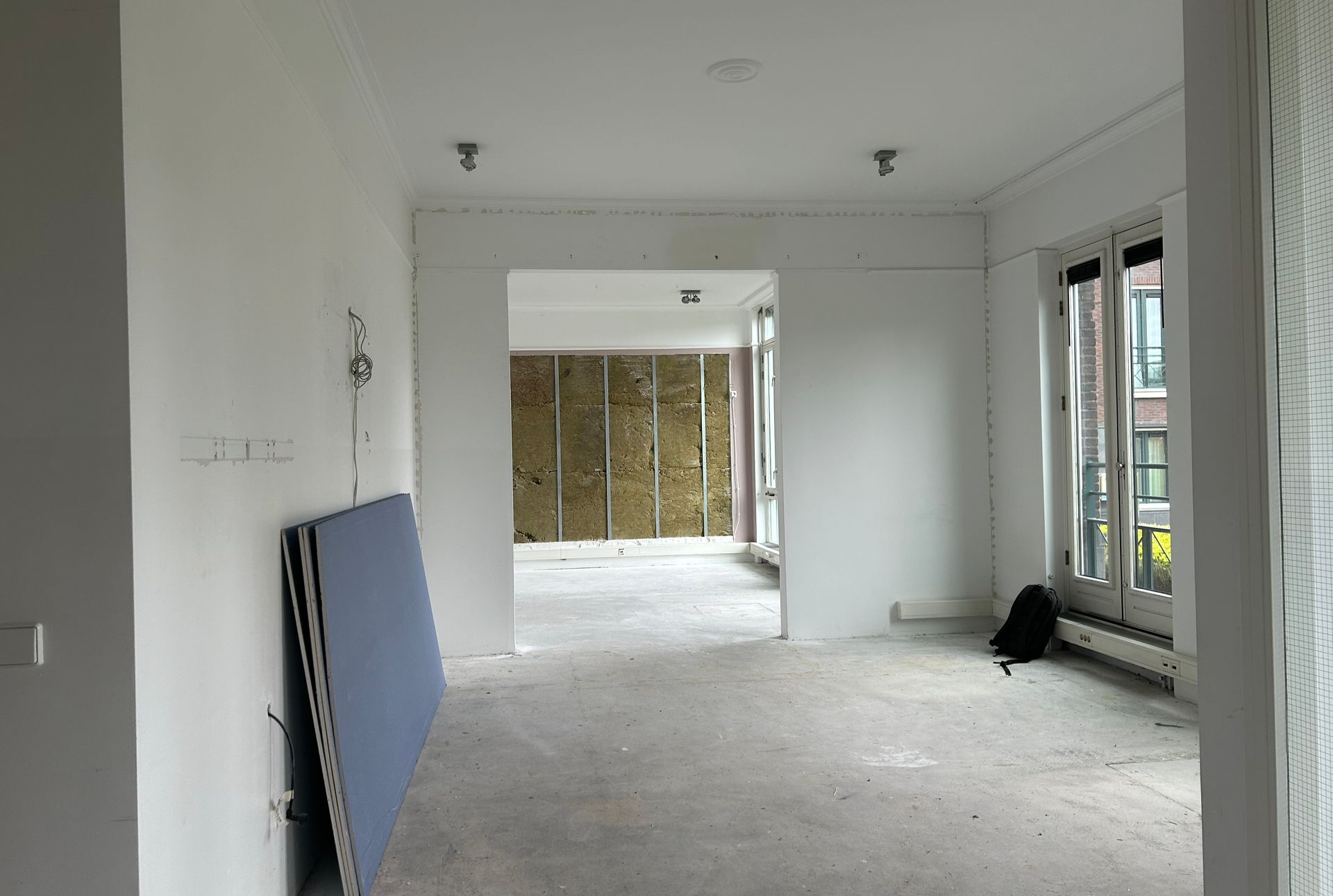 Voor verbouwing: een kamer in renovatie met witte muren, een betonnen vloer, een scheefstaande blauwe gipsplaat en een muurframe aan de achterkant in het kantoor van The Future Group  Interieurontwerp KuóStudio 