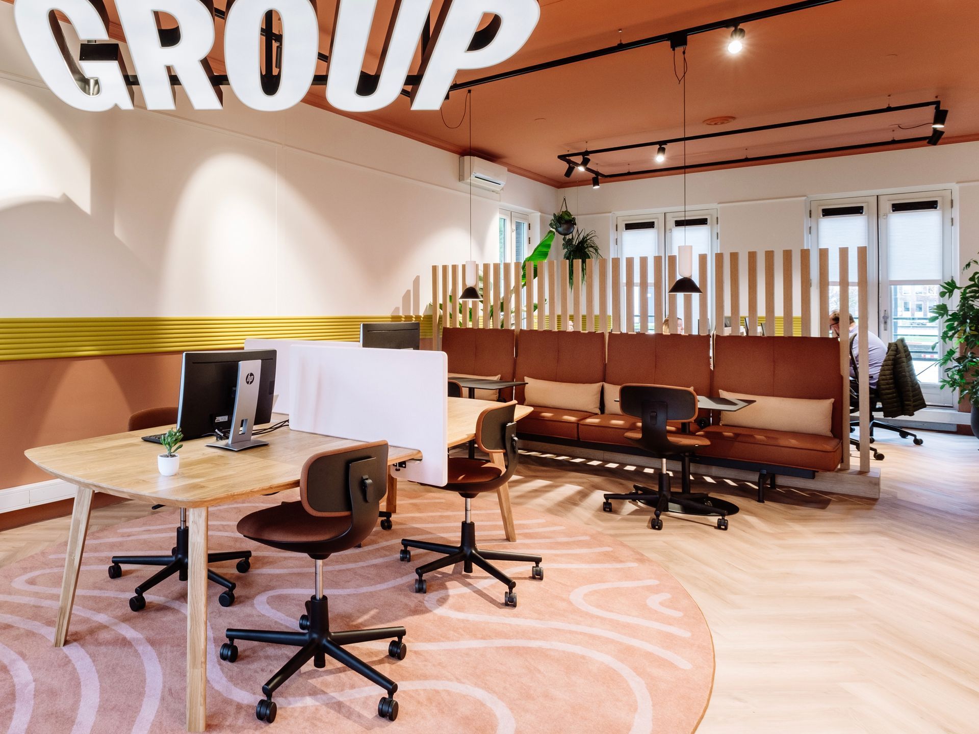 Na verbouwing: Moderne kantoorruimte met een houten bureau, bureaustoelen op wielen en een bruine bank op een roze vloerkleed met patroon onder een gekleurd plafond in het nieuwe kantoor van The Future Group Interieurontwerp KuóStudio