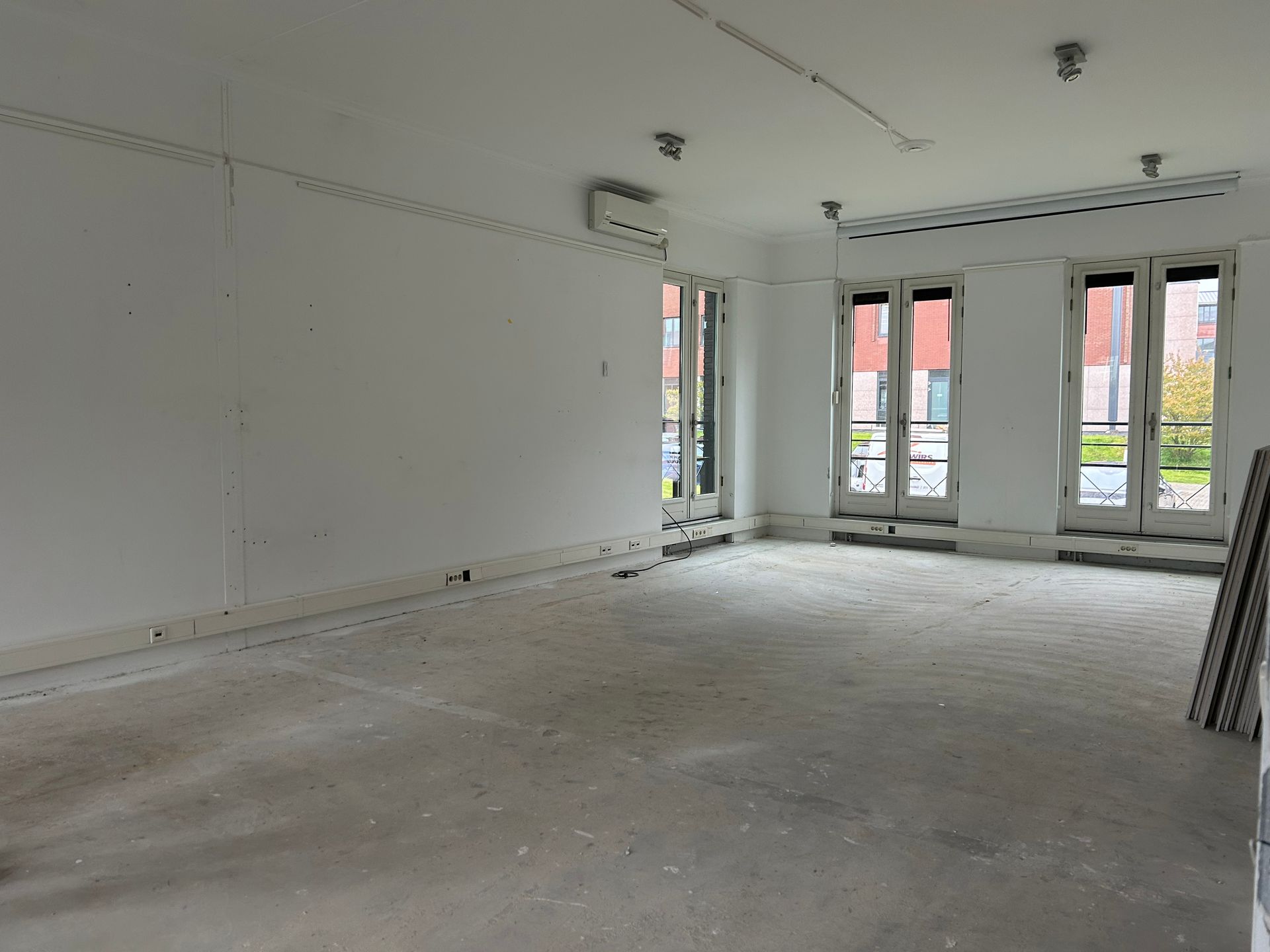 Voor verbouwing: Een lege kamer met witte muren, een betonnen vloer en drie openslaande deuren die natuurlijk licht binnenlaten in het  kantoor van The Future Group 
