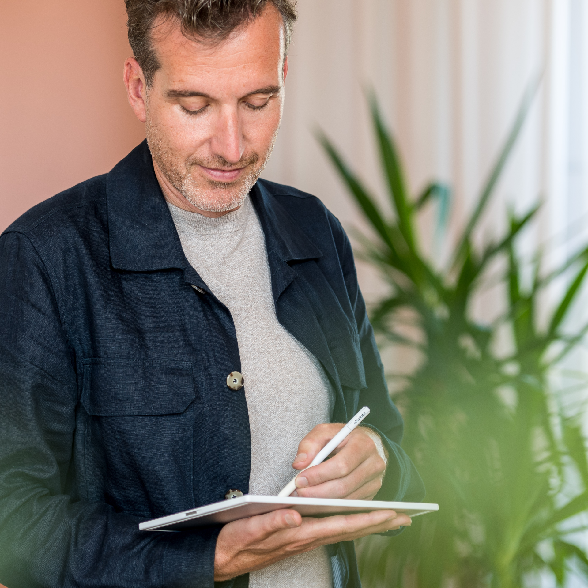 Een persoon, gekleed in een donkere jas over een grijze trui, gebruikt een stylus om op een tablet te schrijven in een lichte kamer met een plant. Implementatie voorbereiding interieurontwerp