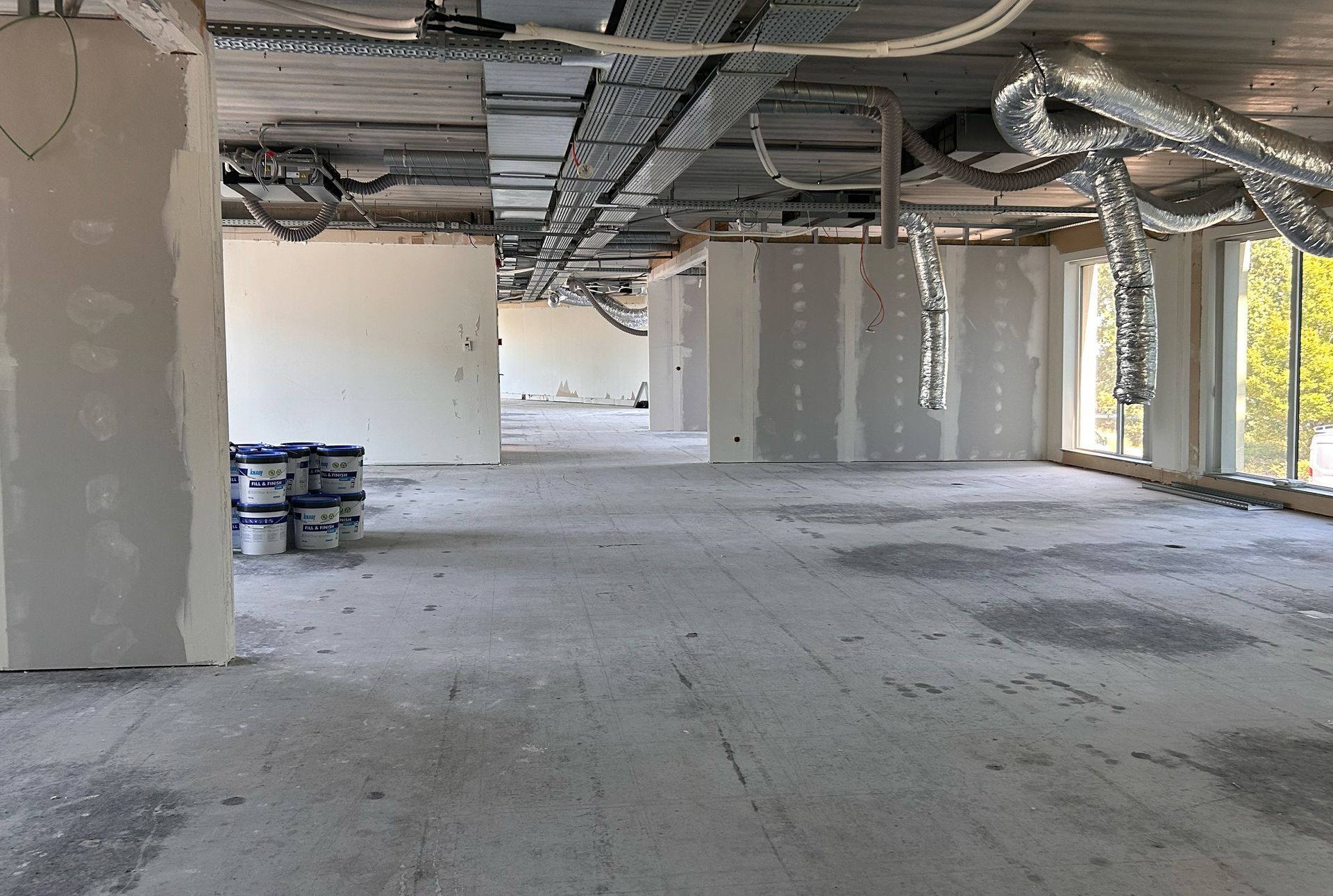 Voor verbouwing: Lege, onafgewerkte binnenruimte met zichtbare plafondleidingen, betonnen vloer en gedeeltelijk in aanbouw zijnde muren.