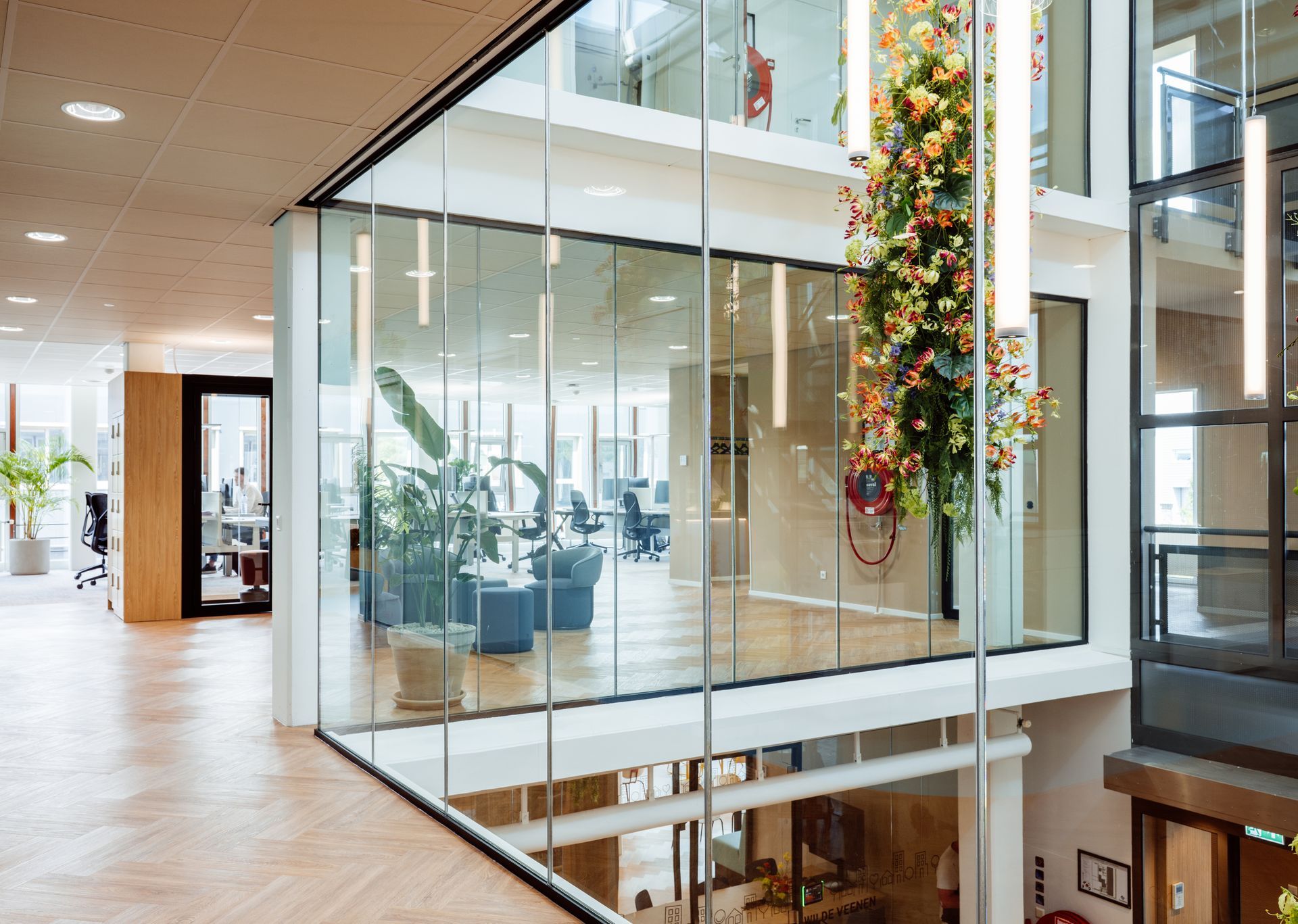 Na verbouwing: Glass-walled atrium with hanging floral decorations and a view into a modern office interior
