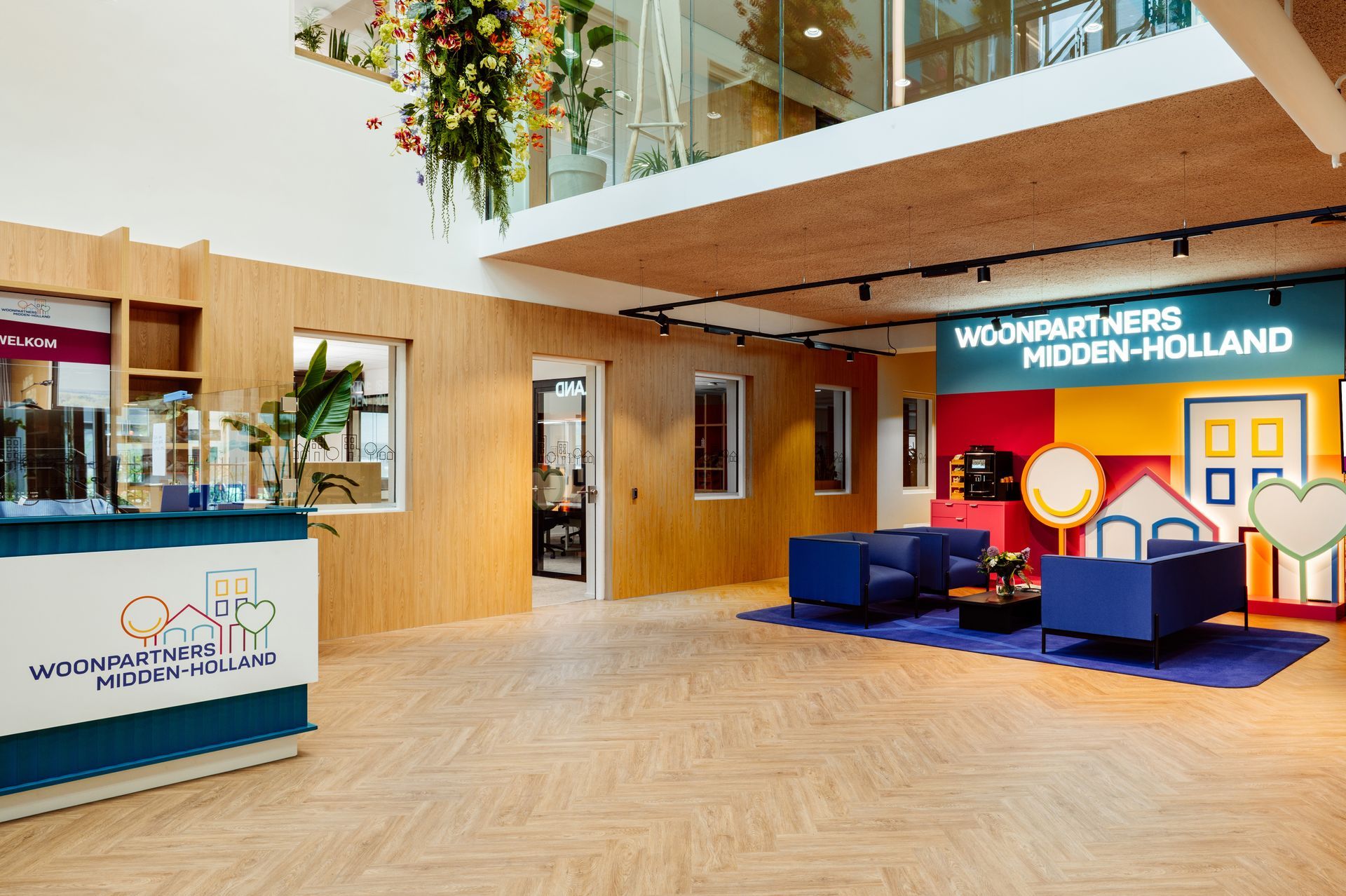 Na verbouwing entree in kantooratrium met een hoge, bloemrijke versiering en zitgelegenheid achter glazen wanden. interieurontwerp door KuóStudio woningcorporatie