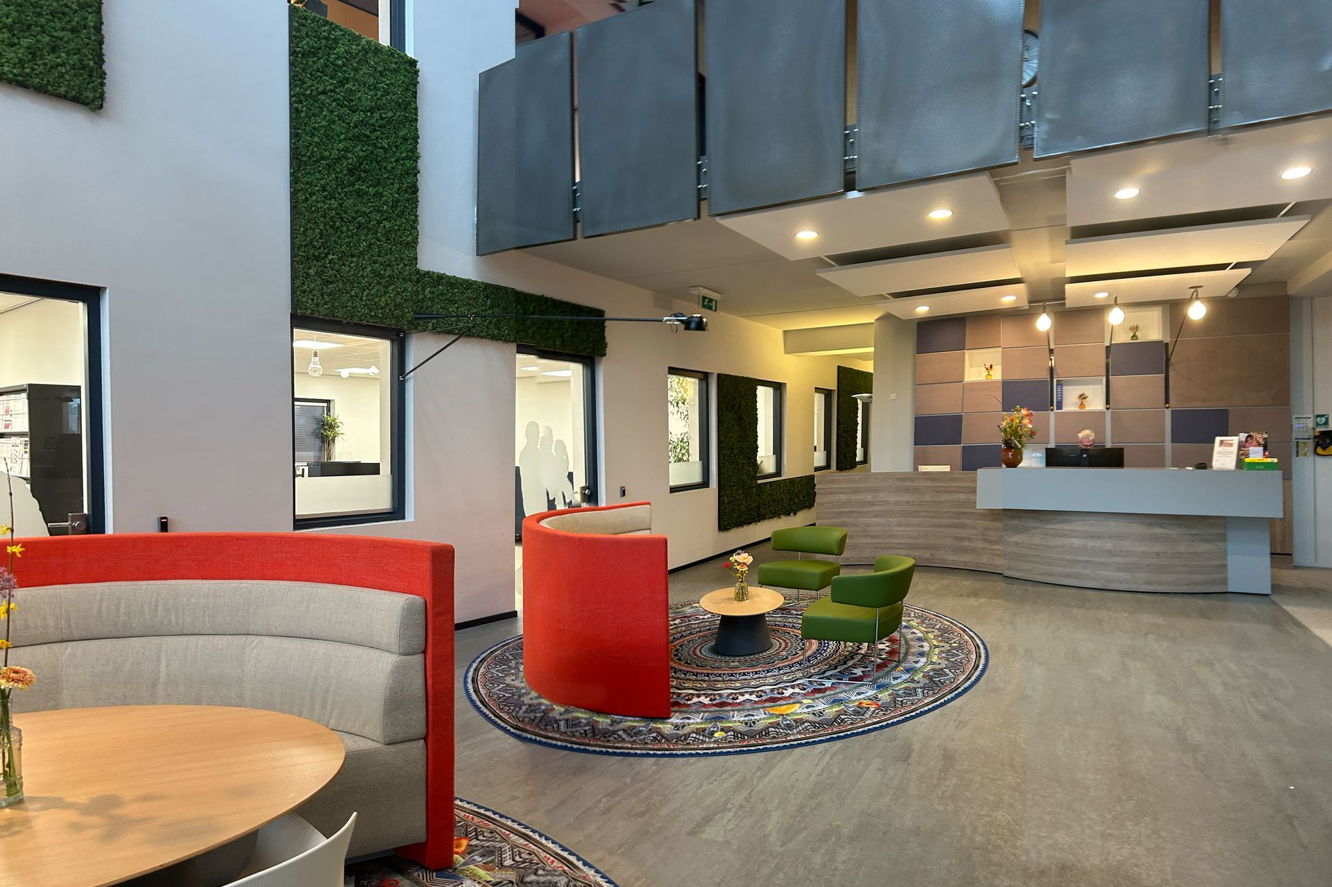 Voor verbouwing gedateerde kantoorlobby met kleurrijke zitmeubels, een receptiebalie en binnenplanten. Woningcorporatie