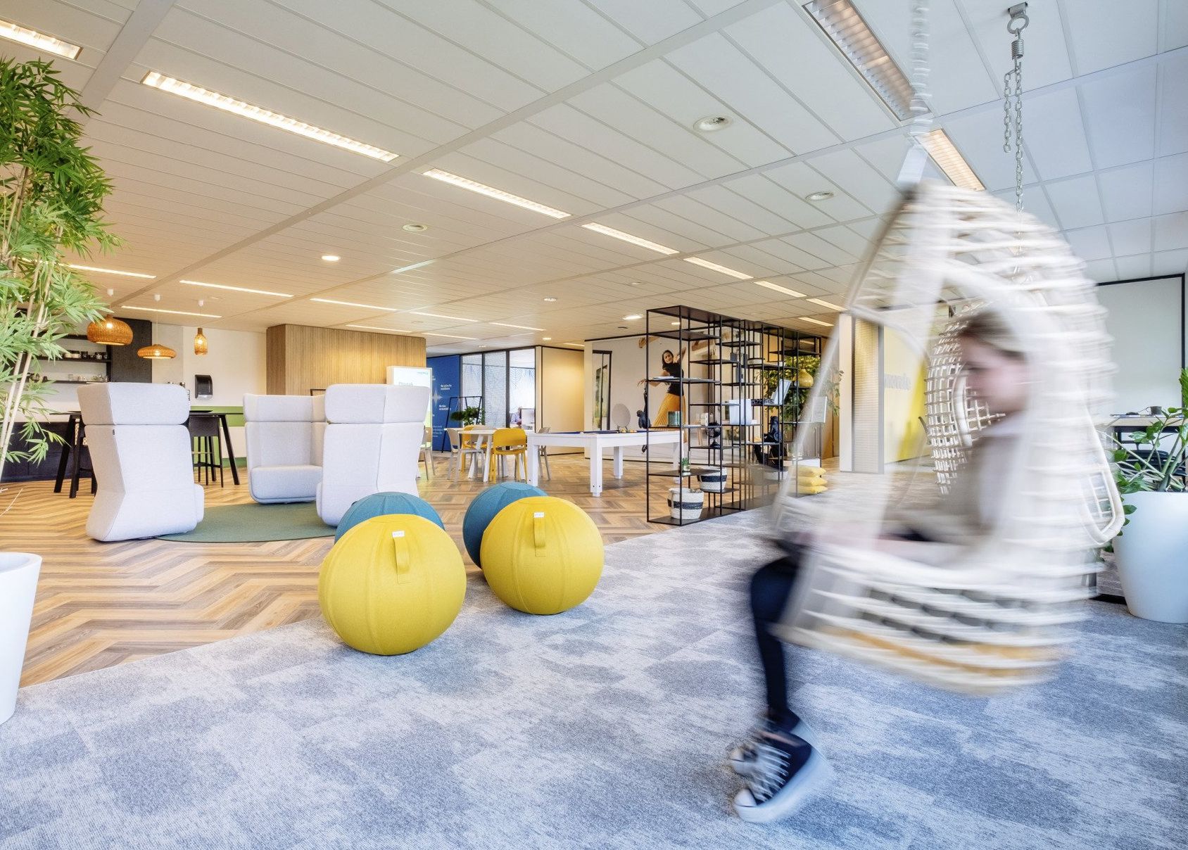 Lichte, moderne kantoorlounge met gele fitnessballen en een wazig figuur in een hangstoel.