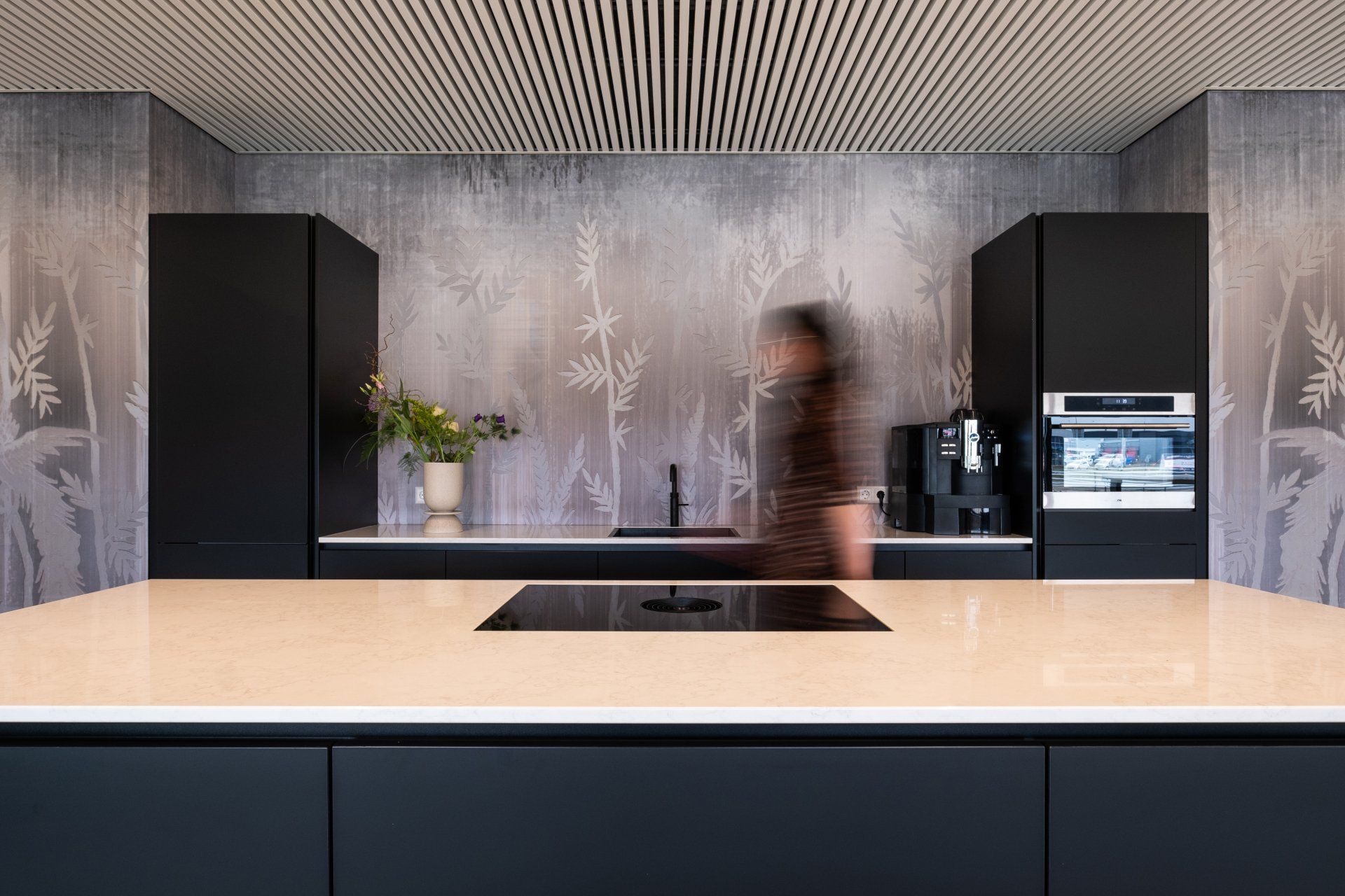 Een moderne keuken met matzwarte kasten, een groot wit kookeiland en een decoratieve muur met behang in natuurmotief in het nieuwe kantoor van EBH Legal in Naaldwijk, interieurontwerp door KuóStudio