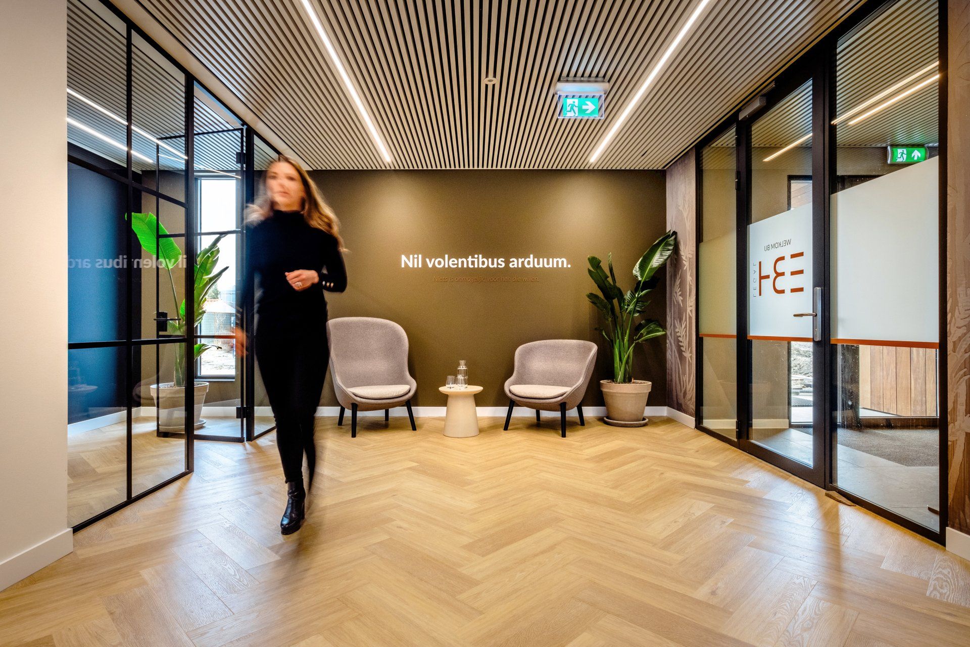 Moderne kantoorlobby met zitgelegenheid, tekst op de muur en een wazige persoon die over de voorgrond loopt.