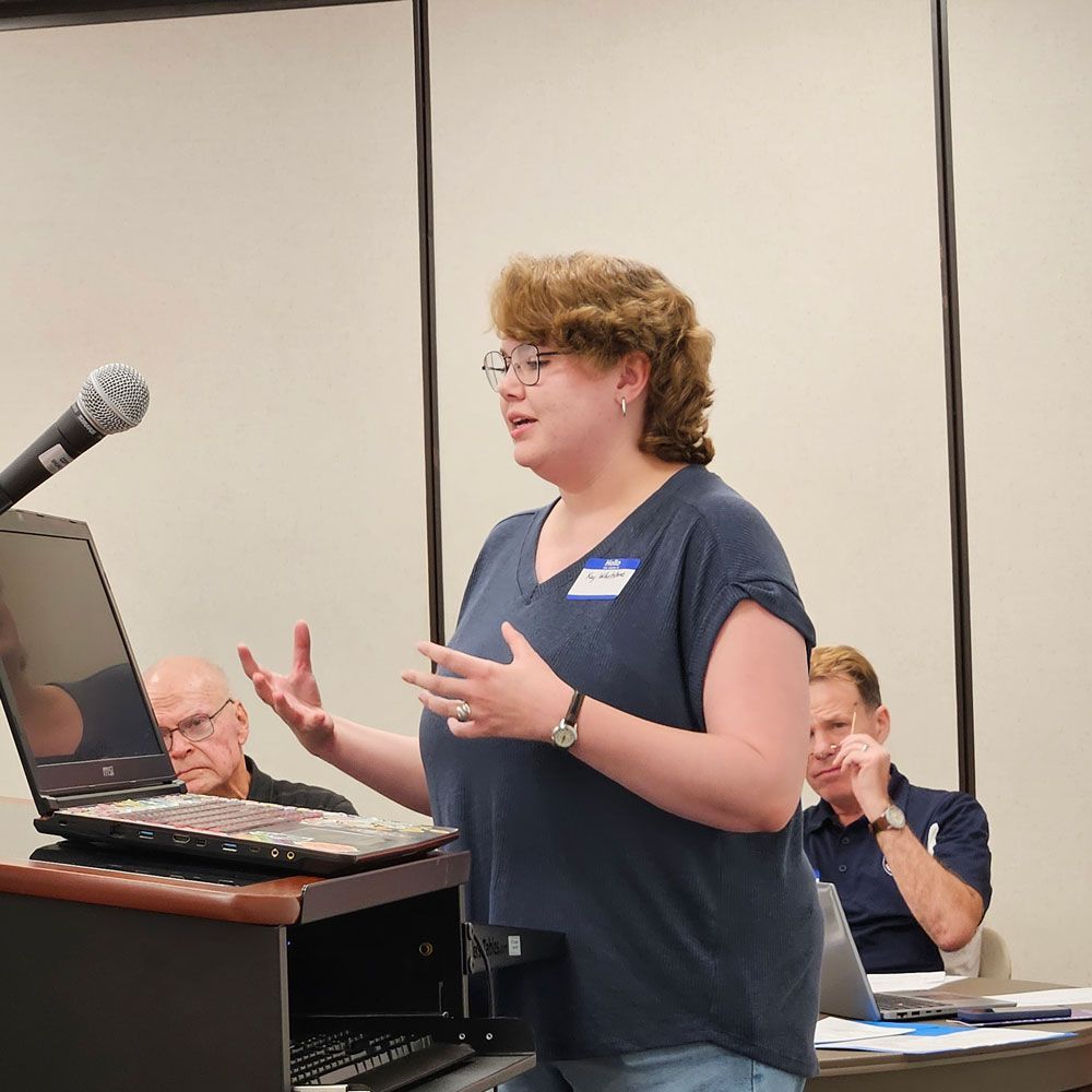 Woman presenting at a podium, gesturing with hands. Two men listen from the background. Inside setting.