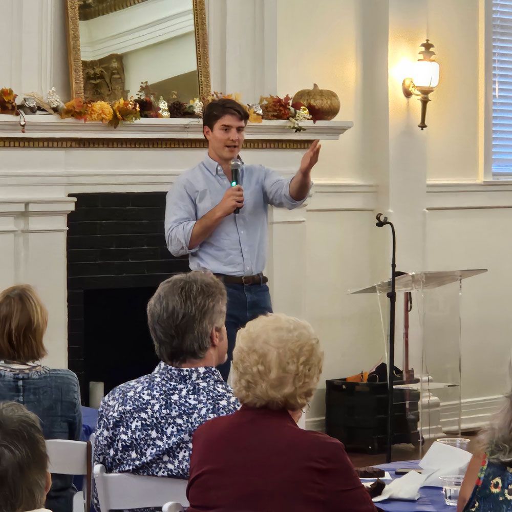 Man speaking at podium, gesturing with right hand; audience seated at tables.