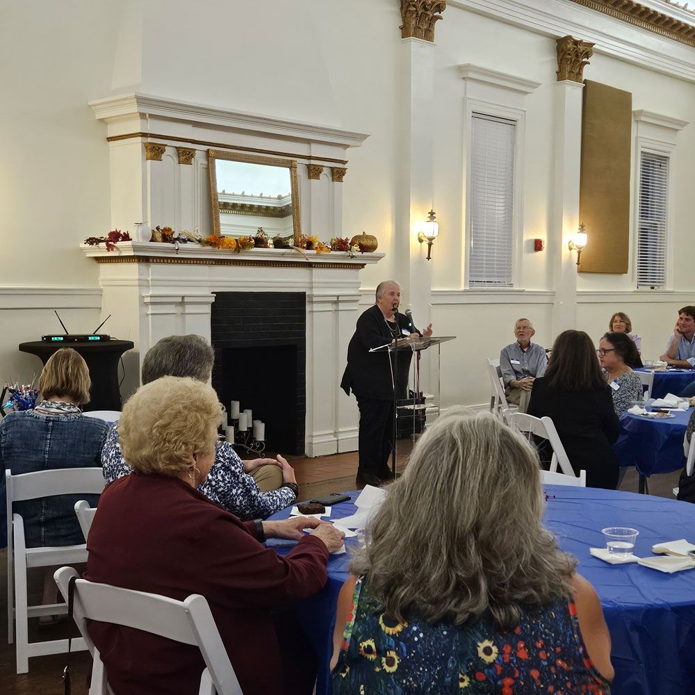 Man speaks to a seated audience at tables covered in blue tablecloths in a large room.