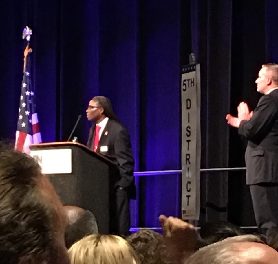 A person in a suit speaks at a podium with an American flag nearby. 