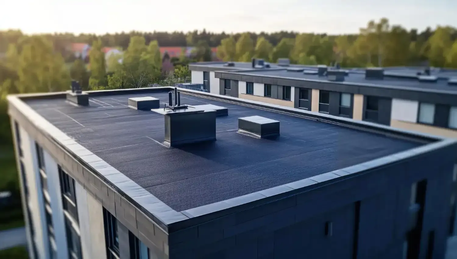 Flat-roof modern building with rooftop vents, seen from above near a tree-lined area