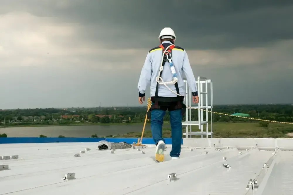 Worker in safety harness walking on a white rooftop under a cloudy sky