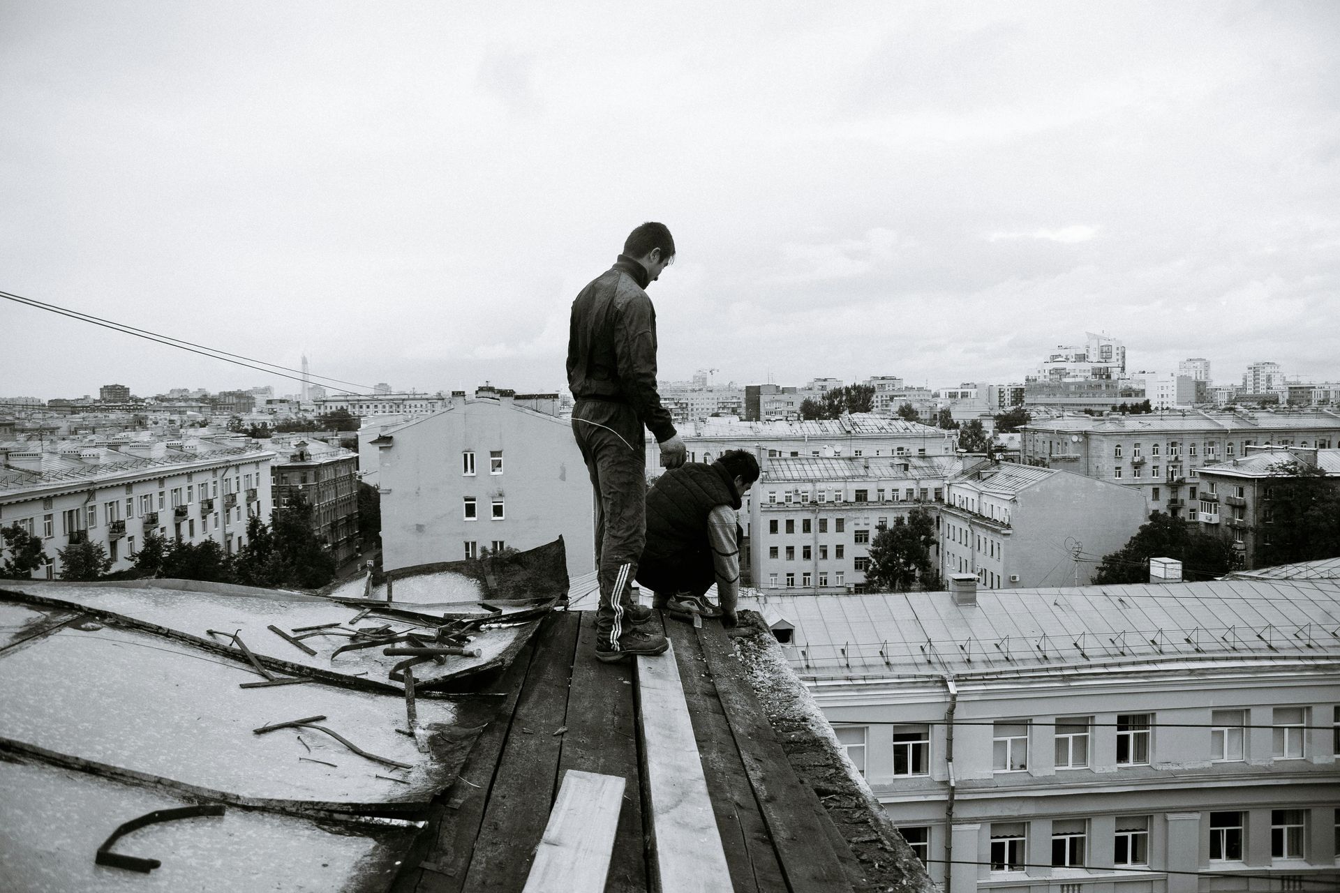 Two people on a rooftop overlooking a cityscape, one standing and one crouching on a narrow beam.