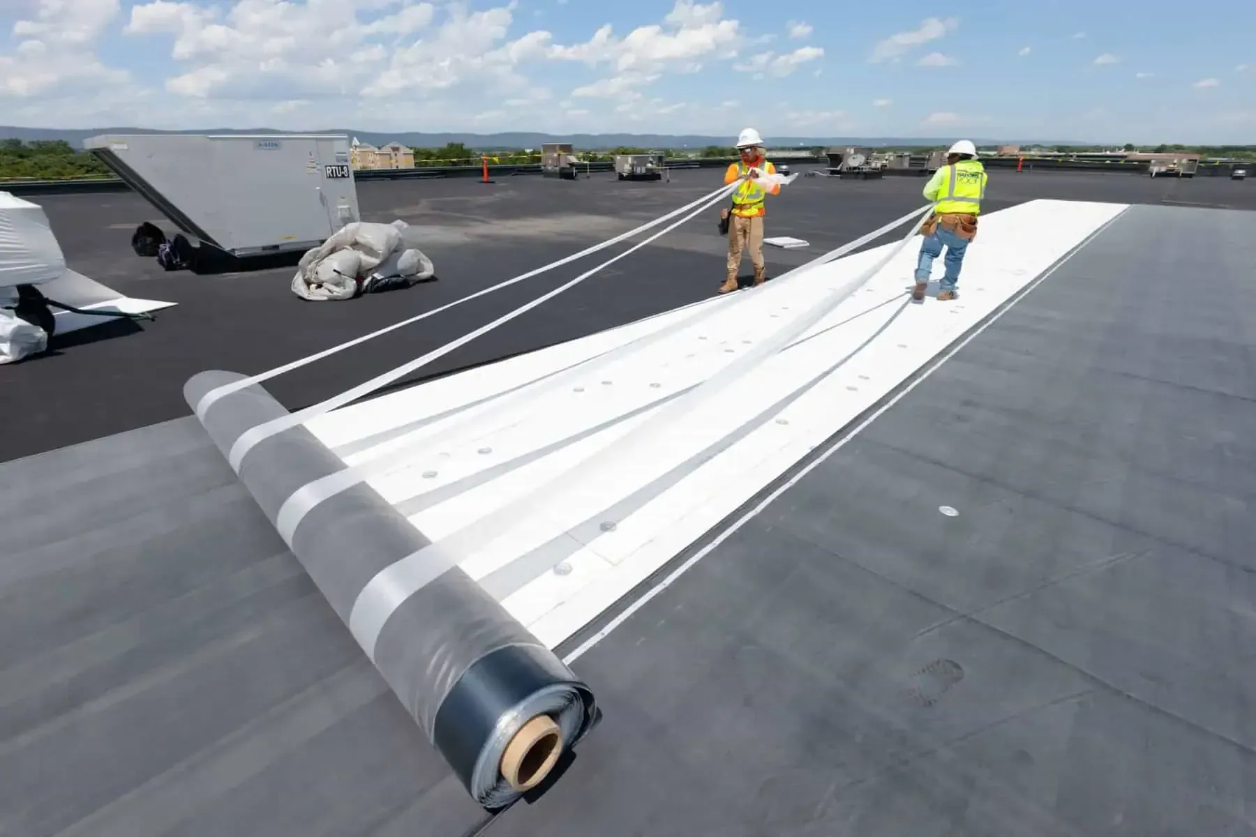 Workers unrolling a large white roof membrane on a flat rooftop under a bright sky