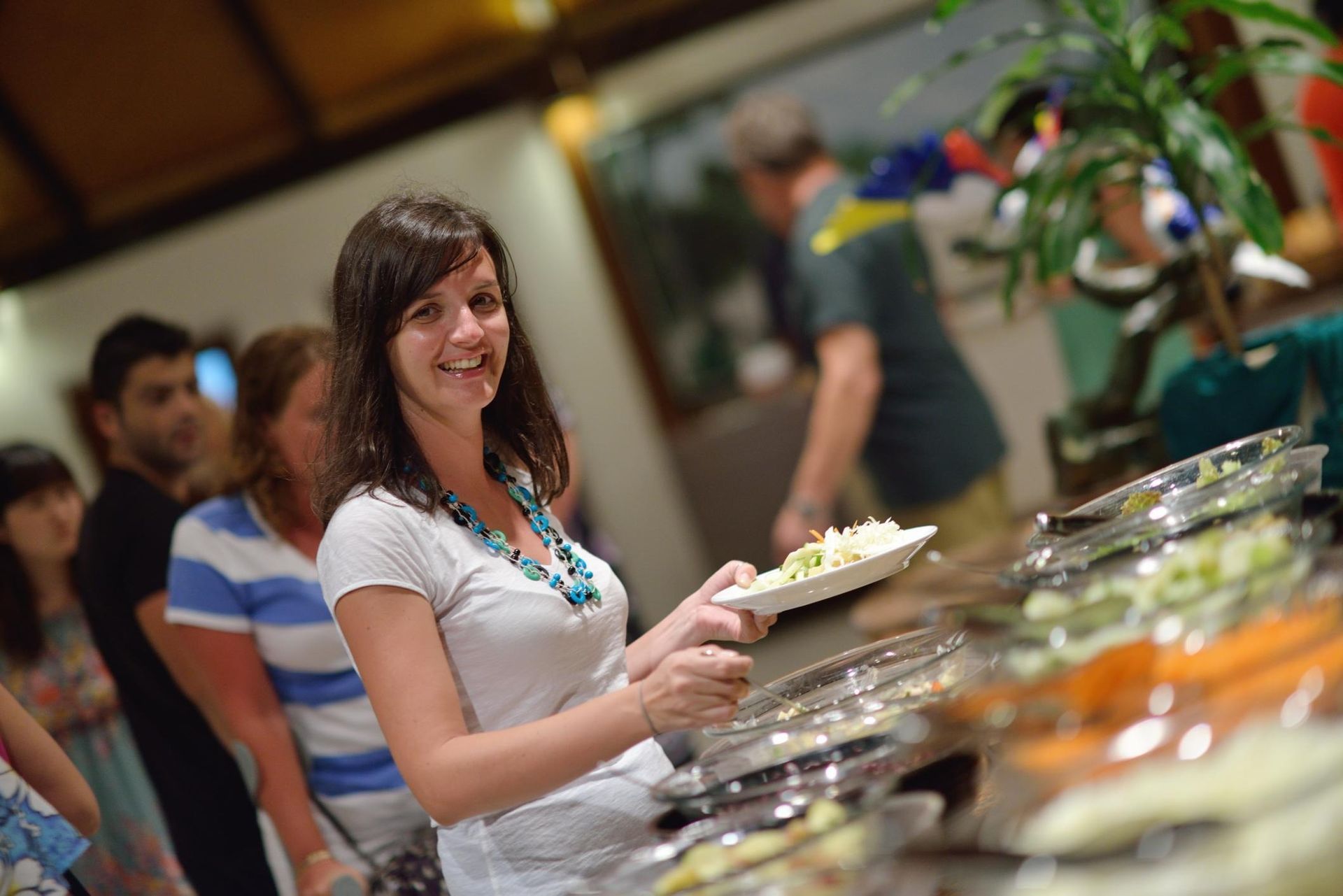 Woman serving herself food at a buffet table, smiling in a bright indoor dining area.