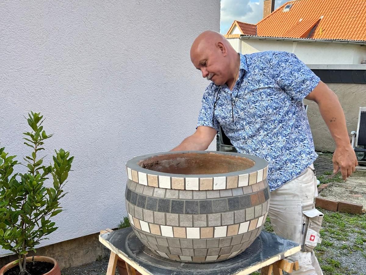 FliesenLukas gestaltet ein rundes Outdoor-Pflanzgefäß mit dekorativen Mosaikfliesen – präzise Handwerksarbeit im Außenbereich