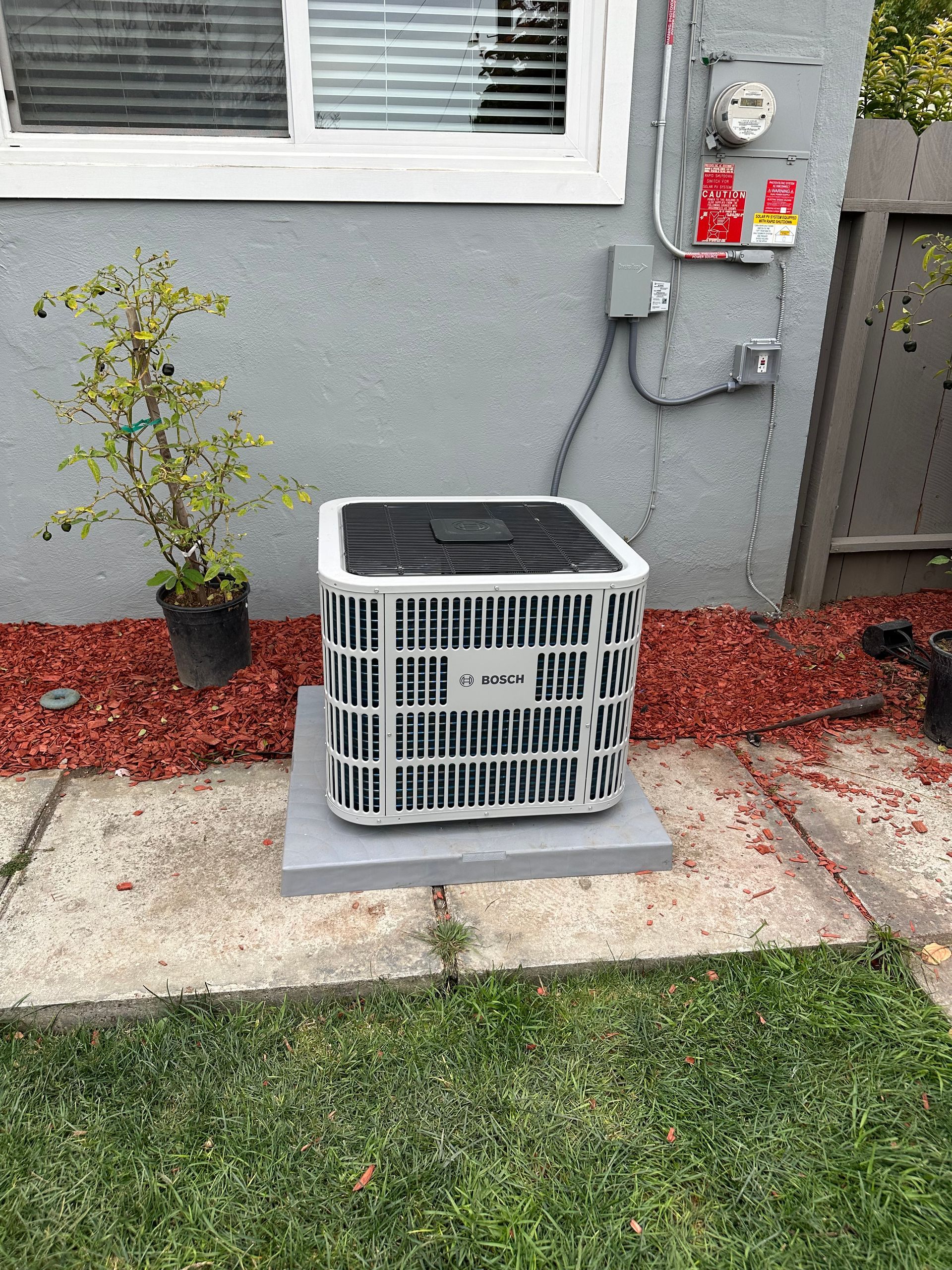 Air conditioning unit on a concrete pad next to a house with red mulch and a small potted plant.