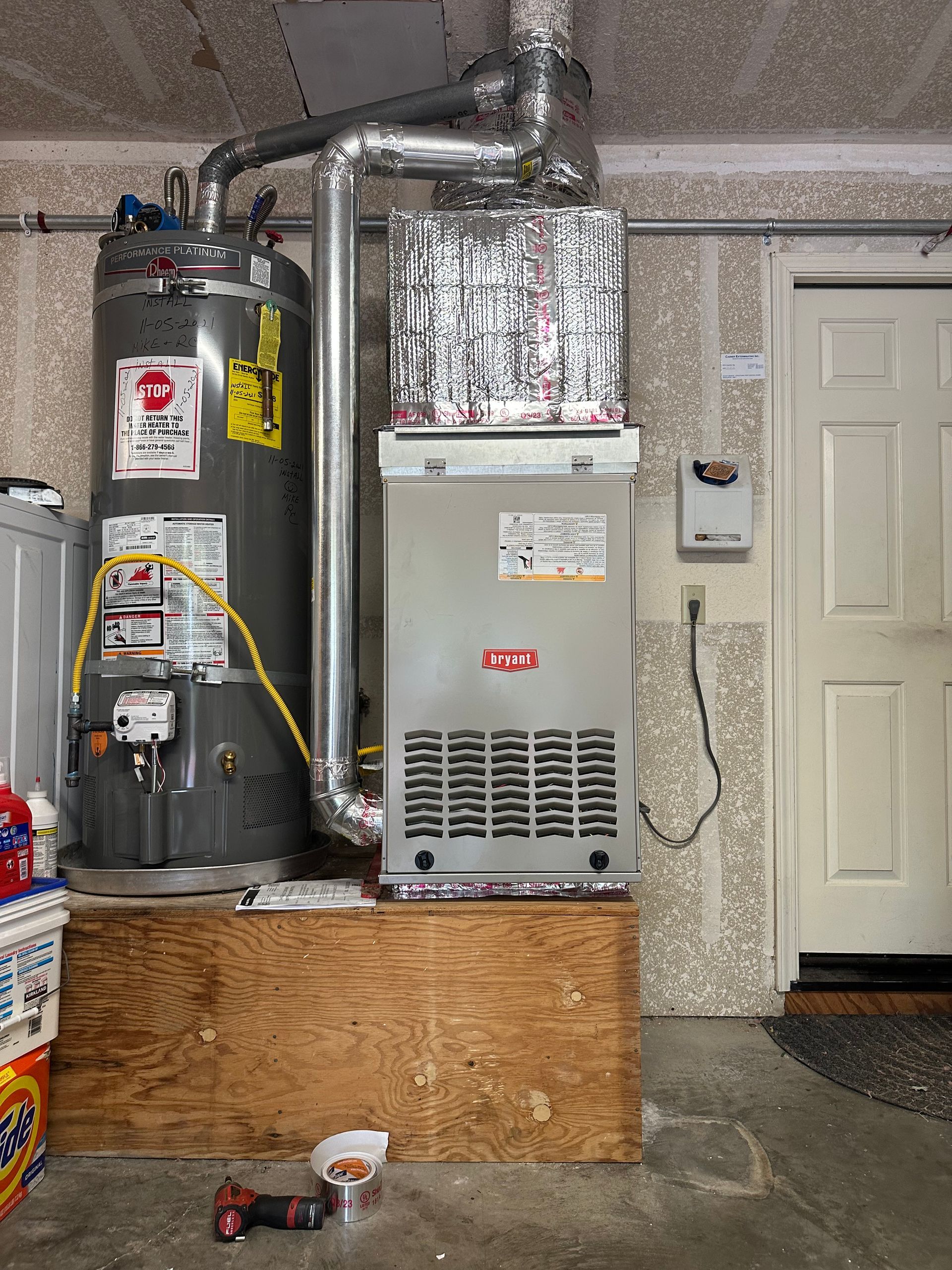 A water heater and furnace unit in a garage, elevated on a wooden platform, next to a white door.