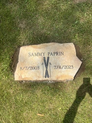 Gravestone of Sammy Paprin, dates 8/7/2003 - 7/4/2023, with crossed skis, in a grassy setting.
