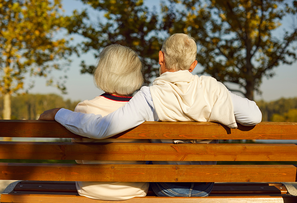 Couple sitting on a park bench, arms around each other, backs to the camera, overlooking a lake.