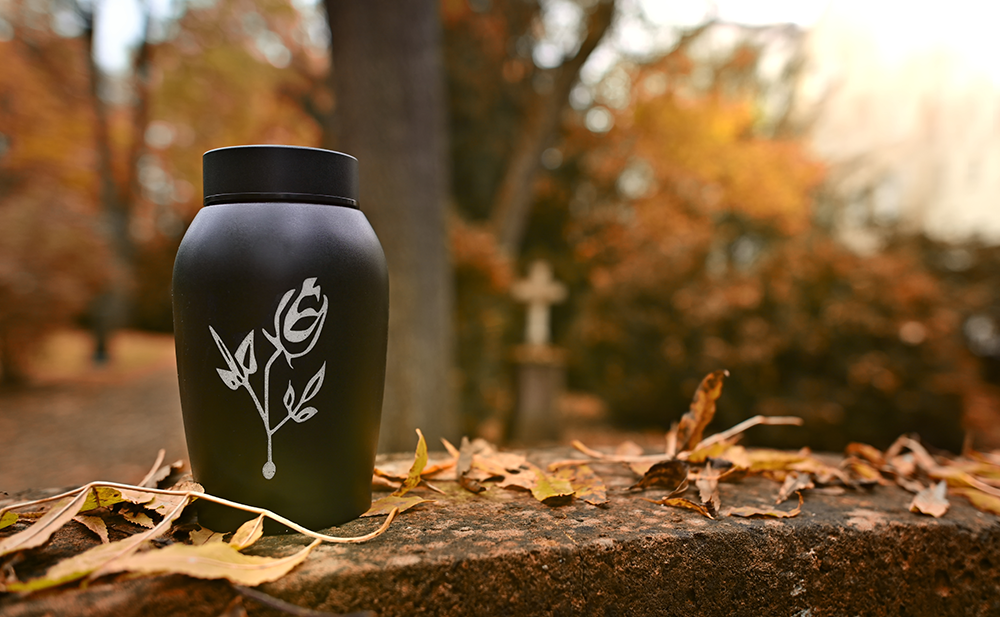Black urn with a rose design, sitting on a stone wall with autumn leaves; trees and a cross in the background.