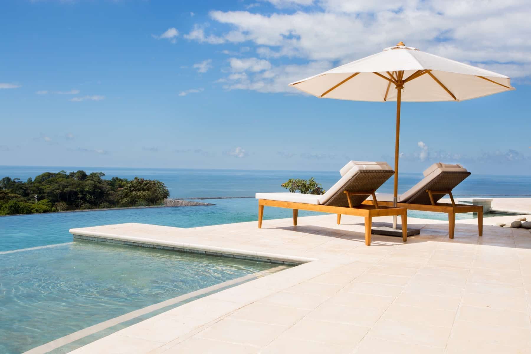 Two lounge chairs under an umbrella next to a swimming pool overlooking the ocean.