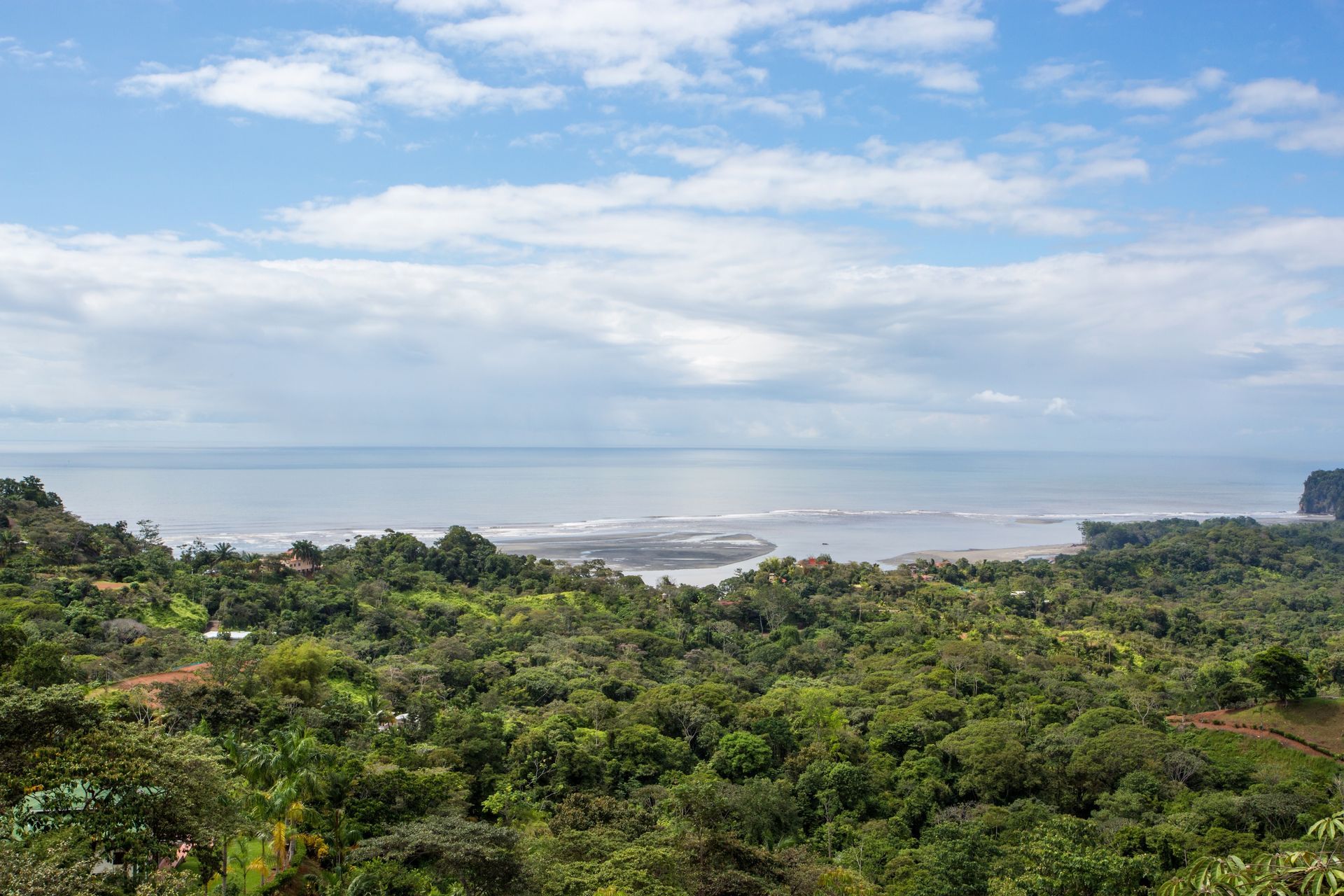 Overlooking lush green forest, a beach, and the ocean under a cloudy blue sky.