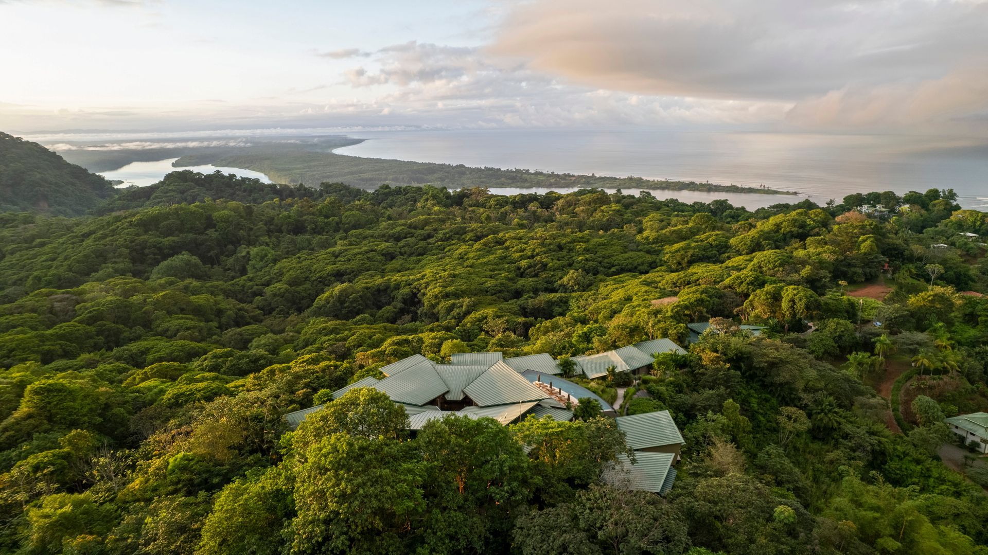 Aerial view of a lush hillside resort with green roofs and winding paths amid tropical forest and mountains