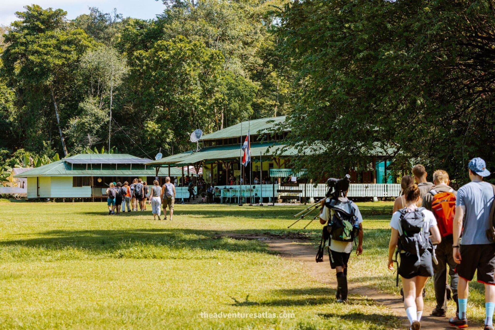 People walking toward a green-roofed lodge in a sunny wooded clearing