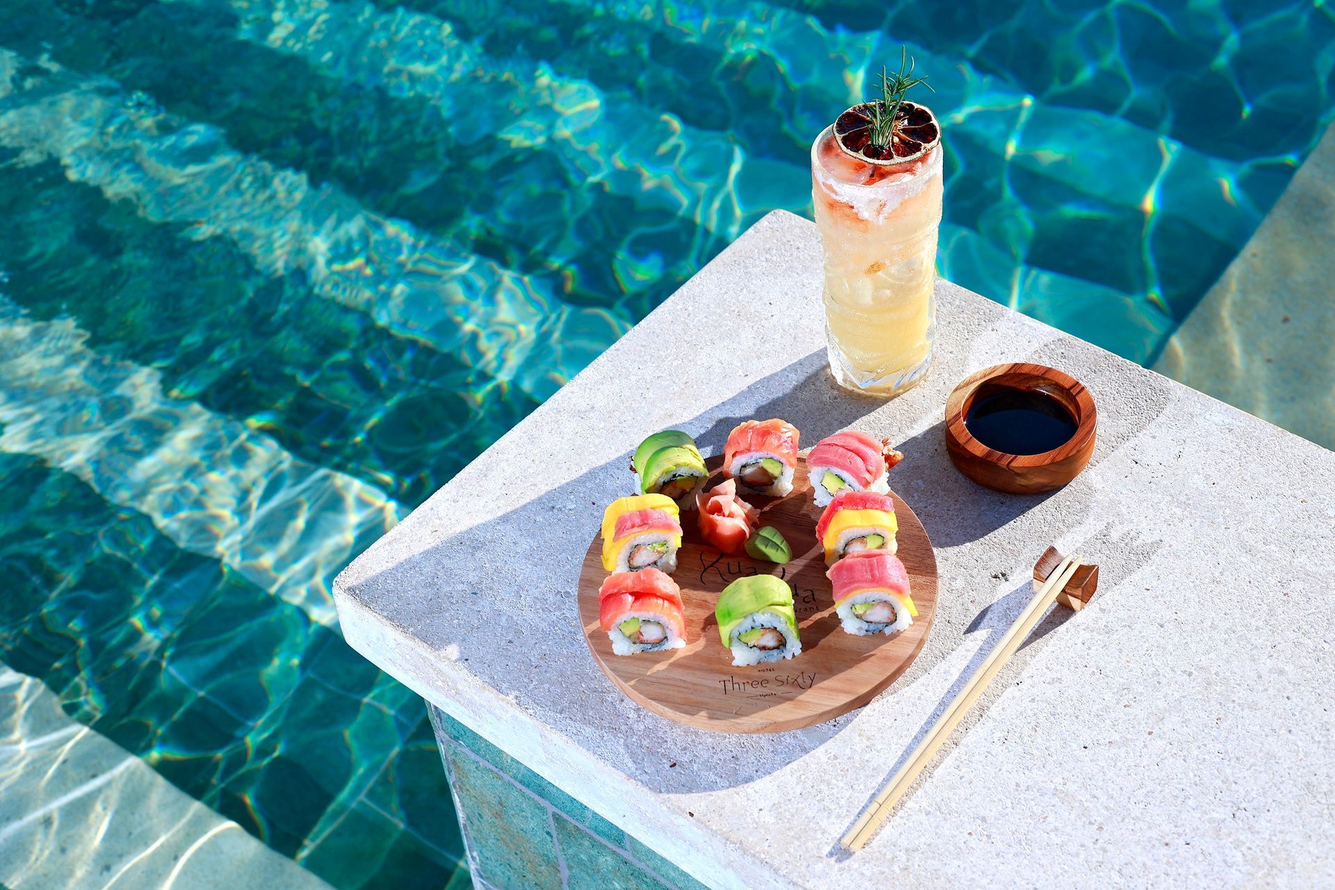 A plate of sushi and a drink on a table next to a swimming pool.