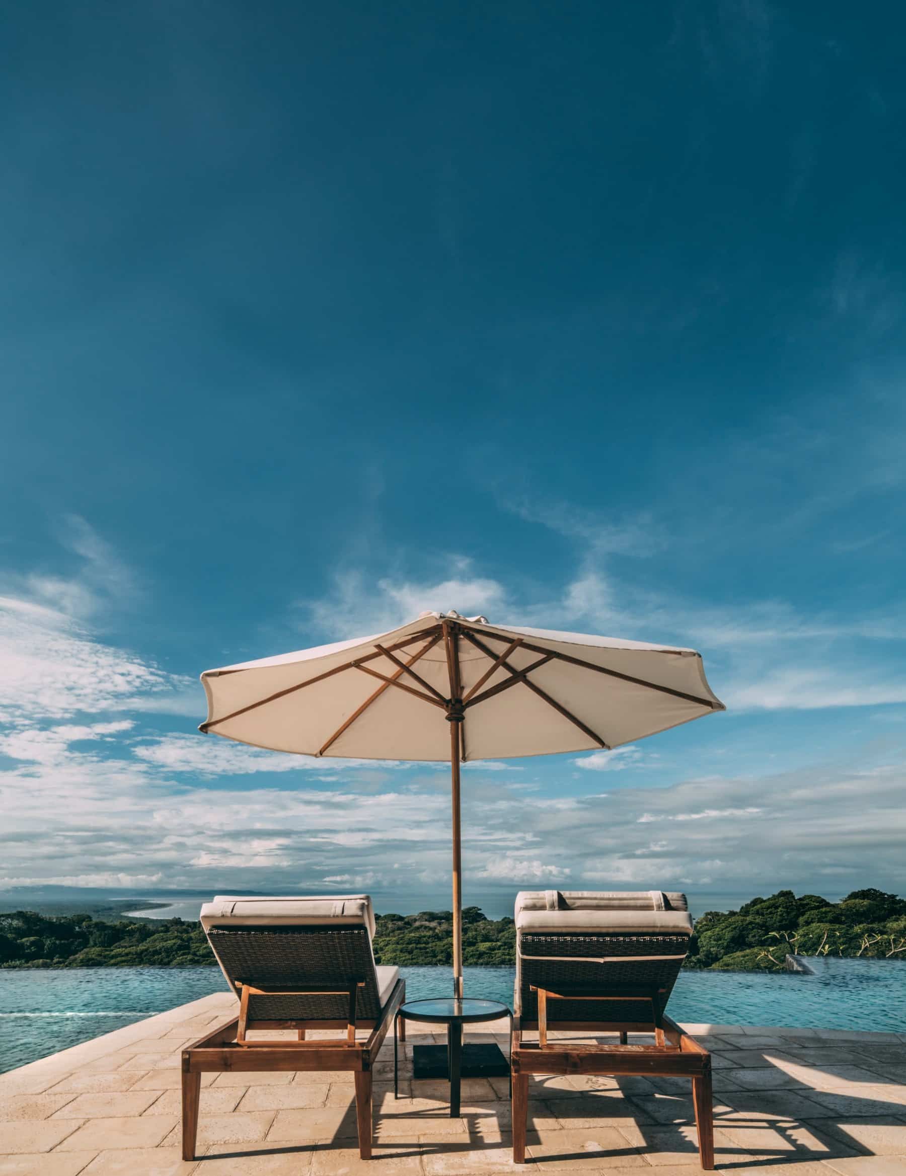 Two lounge chairs under an umbrella next to a swimming pool.
