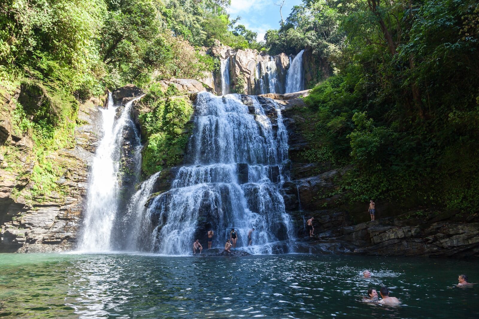 Waterfall cascading into a pool, surrounded by lush green foliage; people swim in the water.