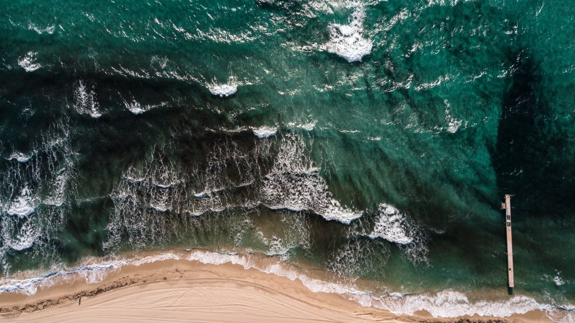 Aerial view of ocean waves crashing onto a sandy beach. Turquoise water, dark shadow, and a wooden pier.