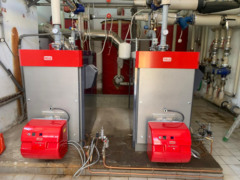 Due caldaie industriali per riscaldamento con bruciatori rossi e pannelli di controllo, installate una accanto all'altra in un locale tecnico.