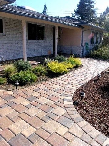 A brick walkway leads to the entrance of a house with a white brick exterior and low-lying shrubs.