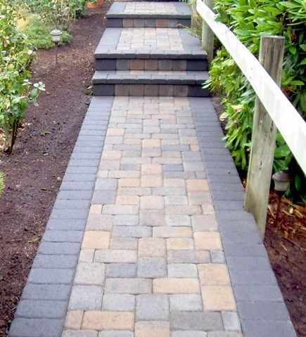 A multi-colored paver walkway with a dark grey border leads to a small set of matching steps flanked by a white fence.