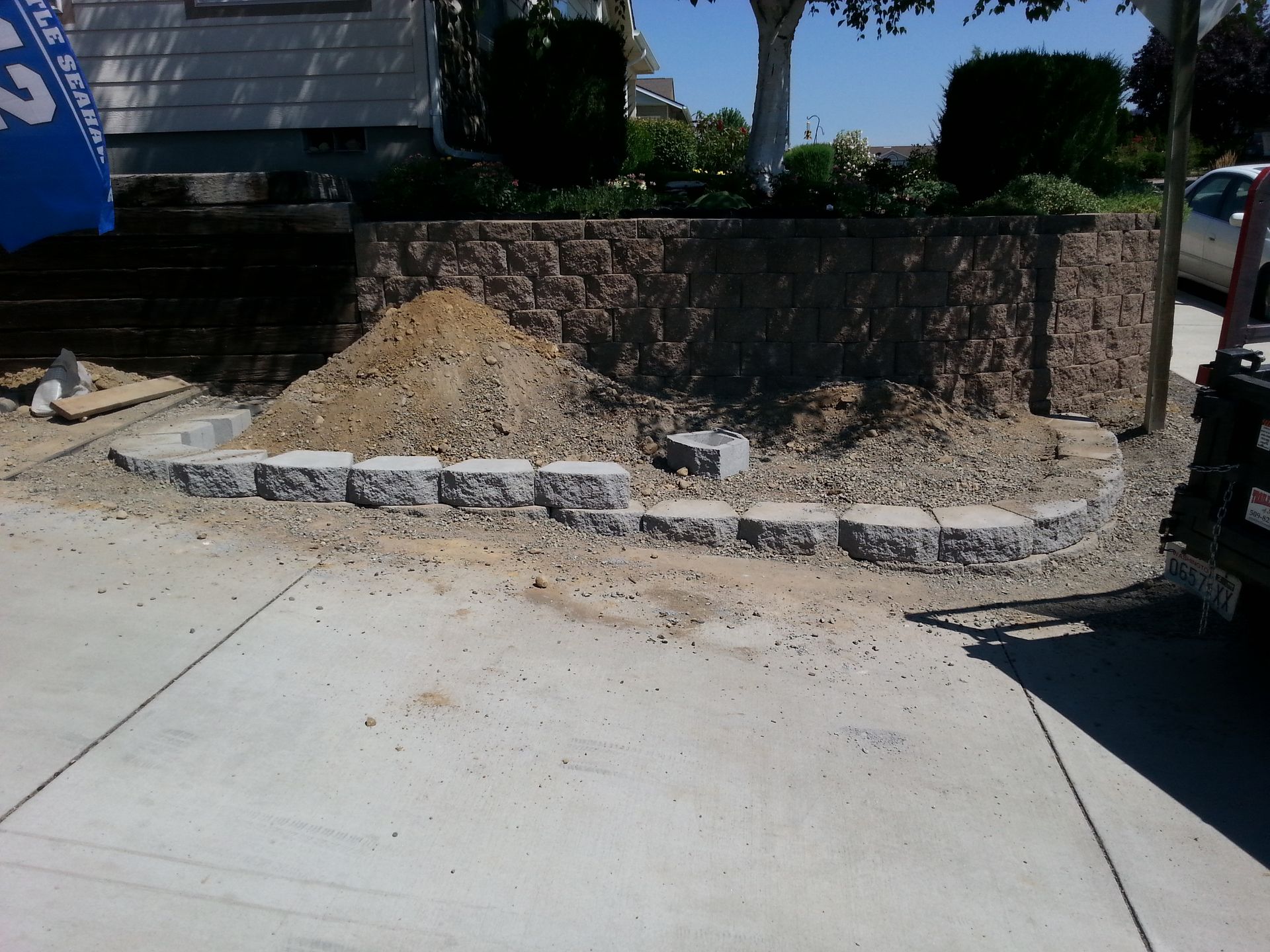 A pile of dirt and gravel sits inside a curved retaining wall made of gray concrete blocks on a residential driveway.