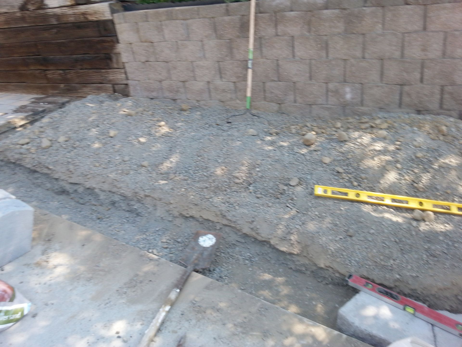 A construction site showing a gravel-filled trench in front of a stone retaining wall, with a level and shovel nearby.