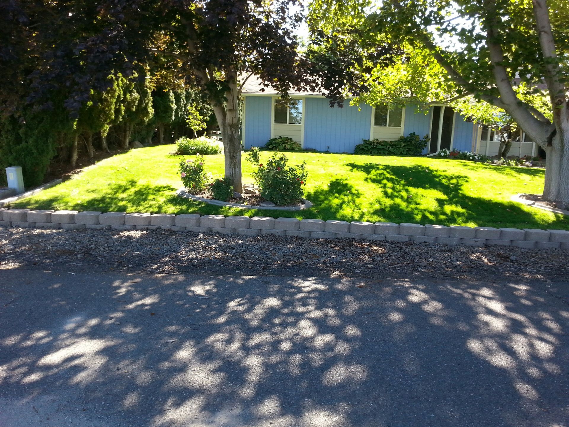 A blue house with a landscaped front lawn, green trees, and a low stone retaining wall next to an asphalt road.
