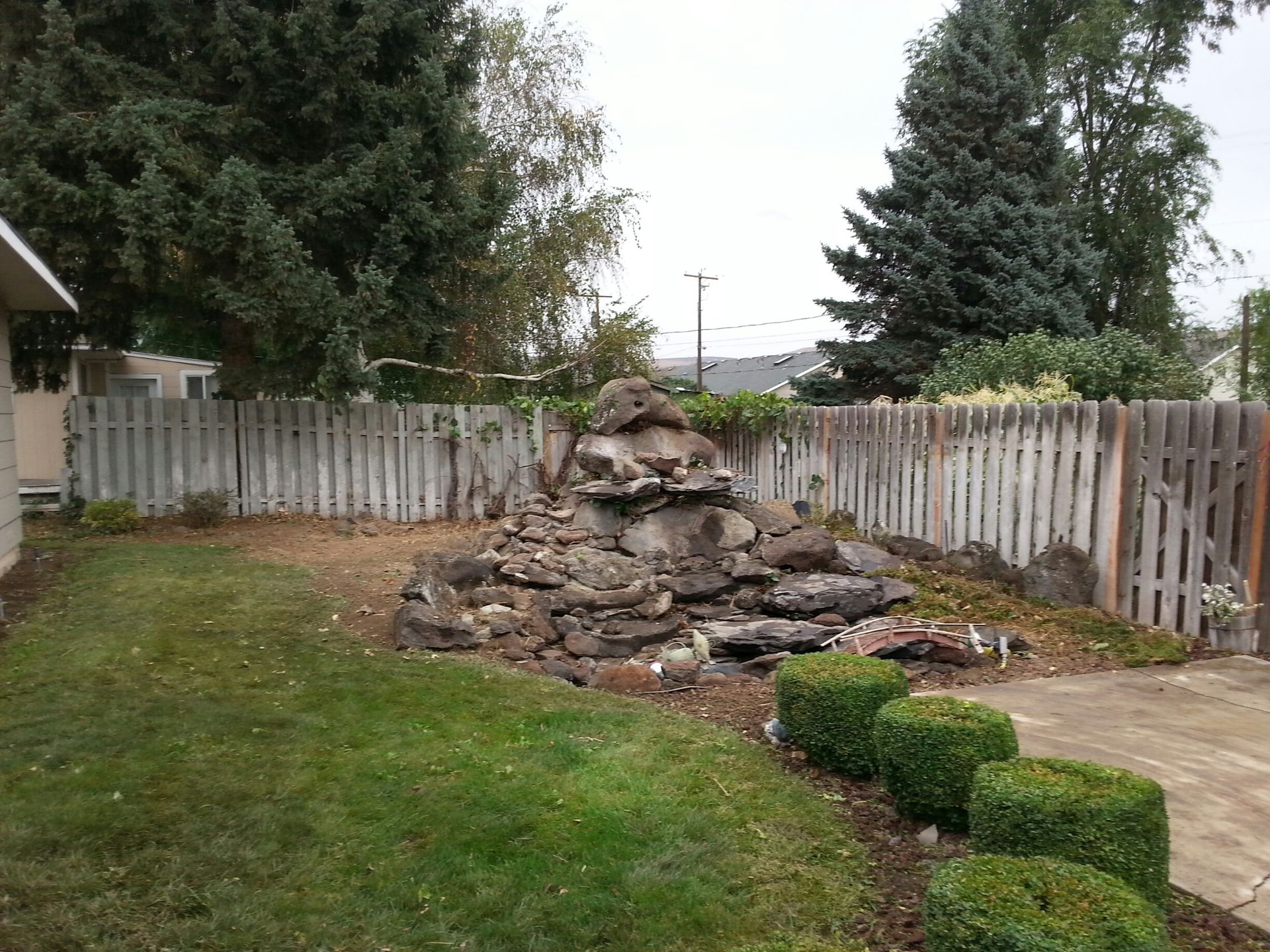 A rock fountain garden feature in a grassy backyard, framed by a light-colored wooden fence and trimmed boxwood bushes.
