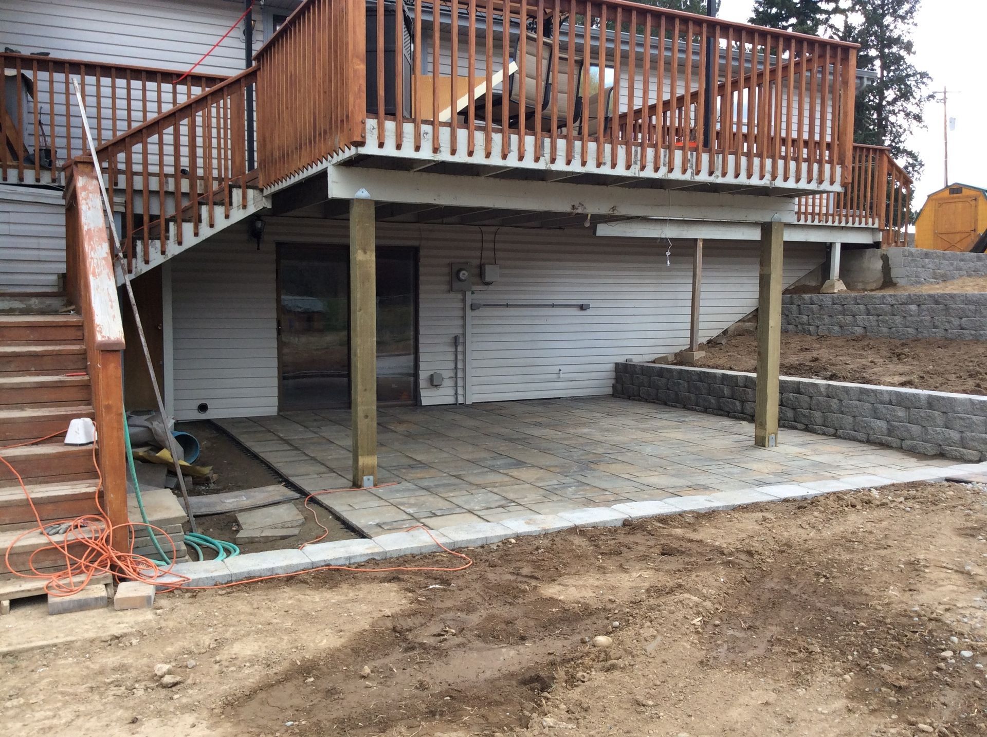 A stone patio sits under a raised wooden deck attached to a house, next to a stone retaining wall and wooden stairs.