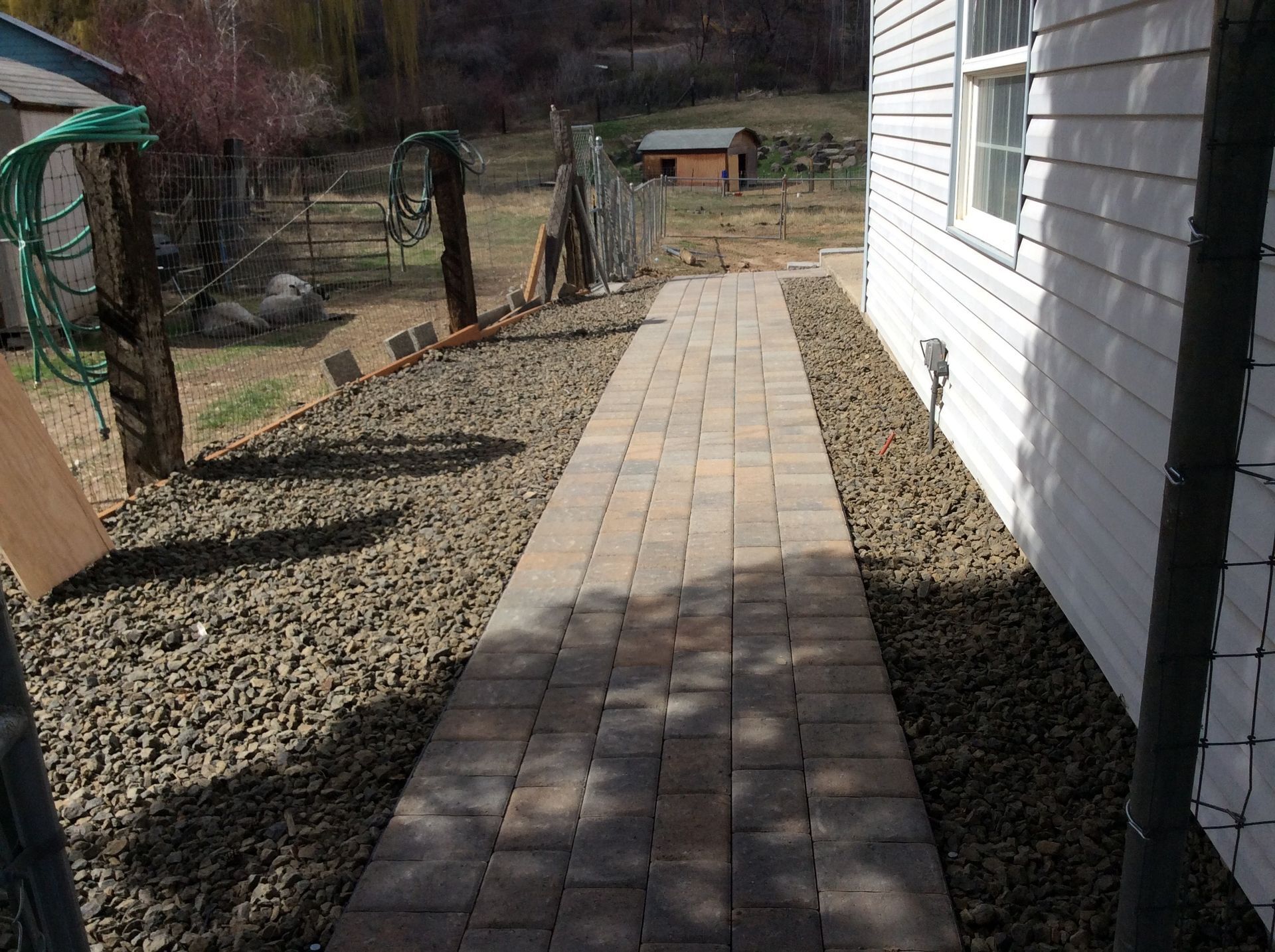 A straight paved stone walkway leads along the side of a white house with gravel ground on both sides.